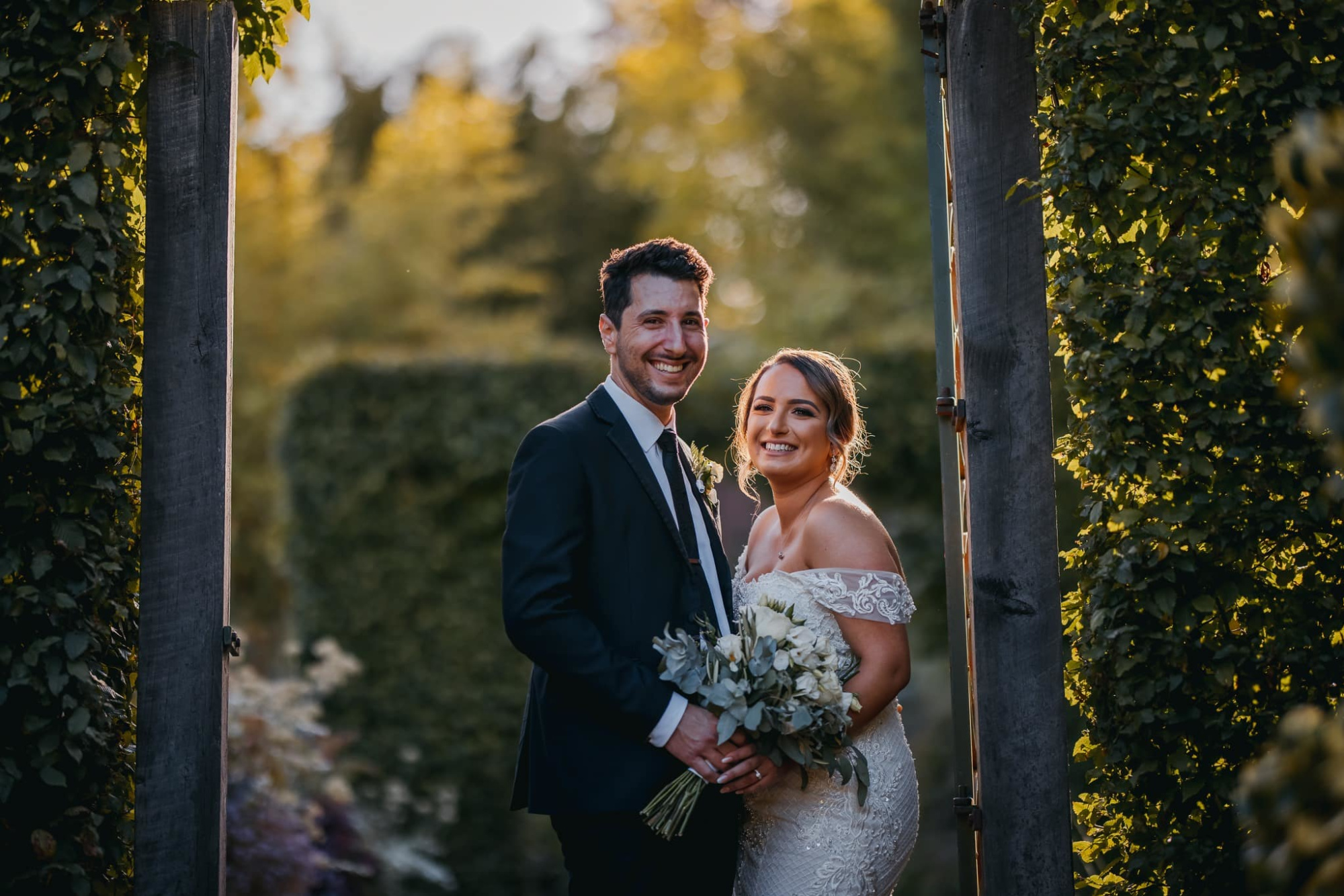 Smiling bride and groom pose with bouquet under a leafy garden arch at sunset.