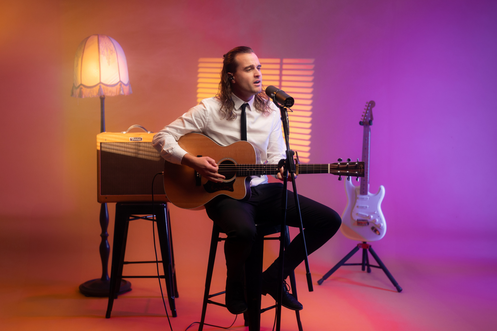 Solo guitarist in shirt and tie performs under colorful studio lighting with microphone and guitars.