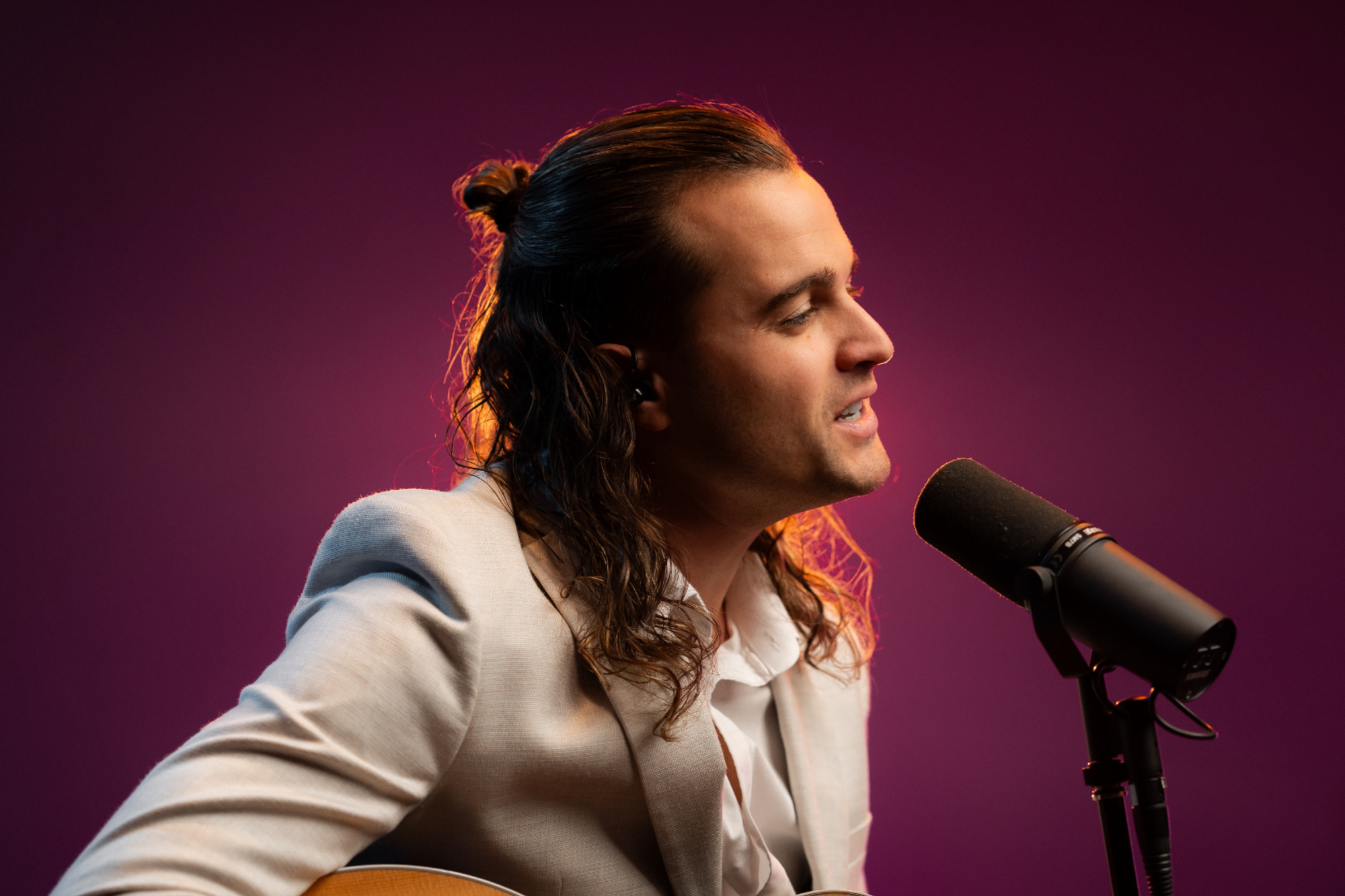 Male musician in a light suit singing into a microphone with guitar against a purple background.