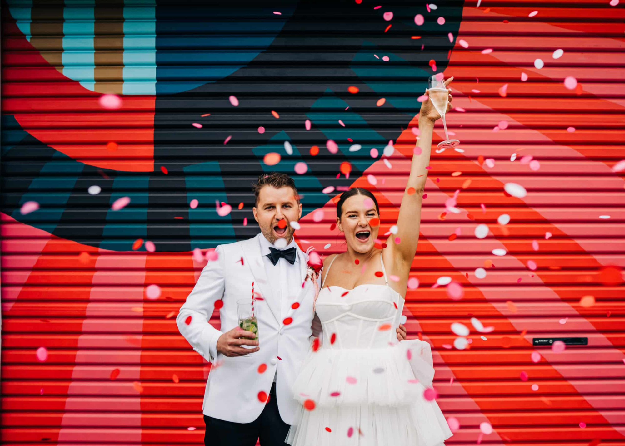 Joyful wedding couple in white outfits celebrating with confetti in front of a colorful striped mural backdrop.