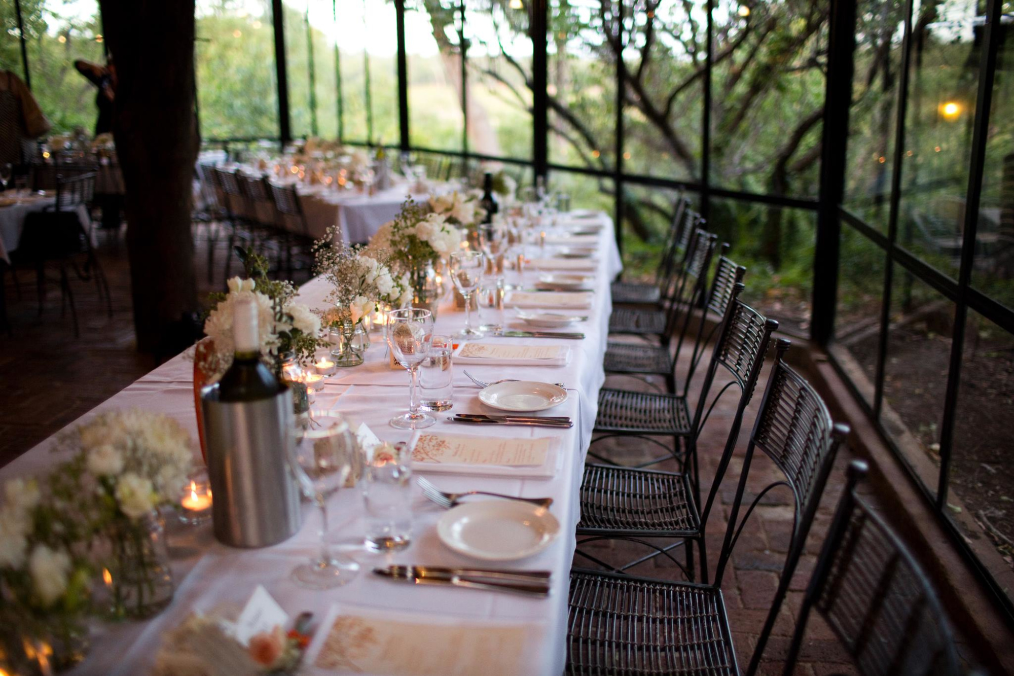 Elegant candlelit wedding reception table set with white linens, floral centerpieces, and glassware in a garden-style venue.