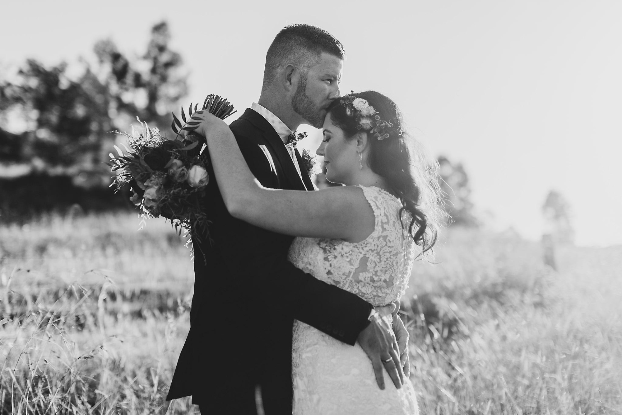 A groom kisses his bride’s forehead as they embrace in a sunlit field holding a bouquet.