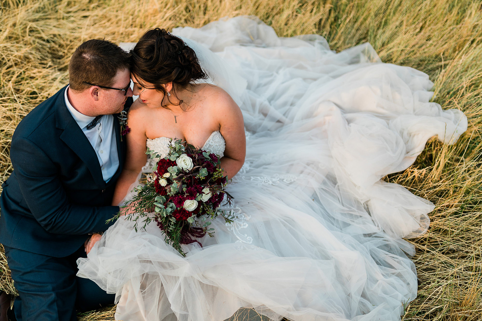 Bride and groom sit close together in a field, her tulle gown spread out around them as she holds a deep red and white bouquet.