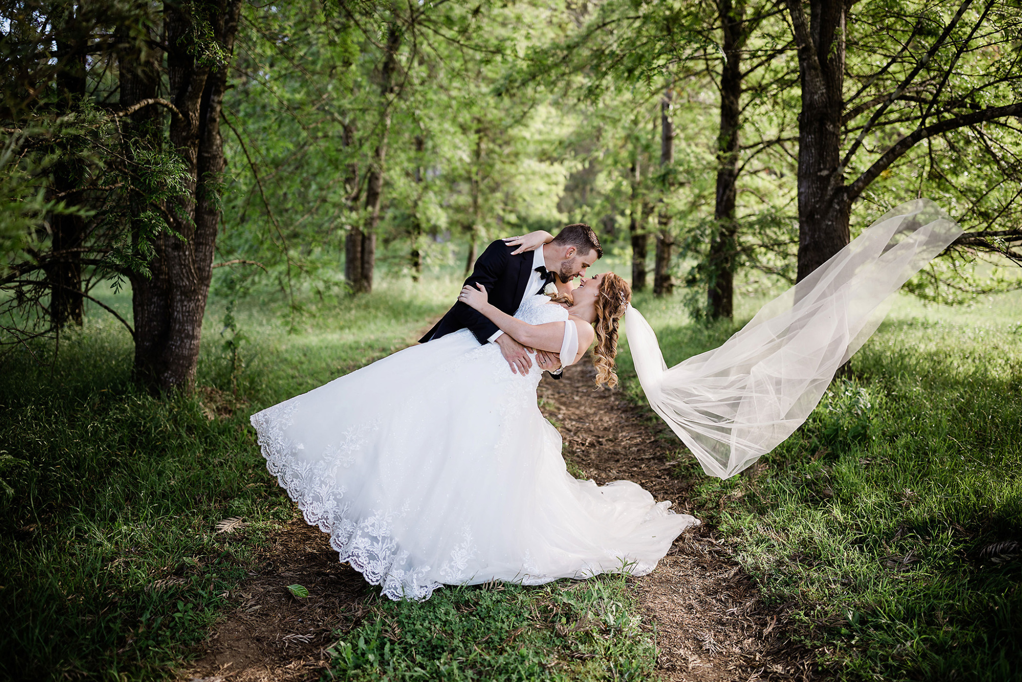 Bride and groom share a romantic dip and kiss on a wooded path, her veil flowing behind in the forest light.