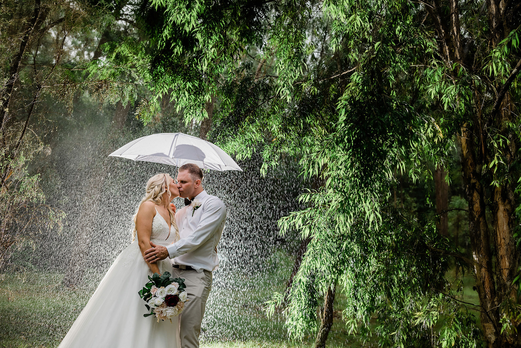 Bride and groom share a kiss under an umbrella in the rain surrounded by lush green trees.