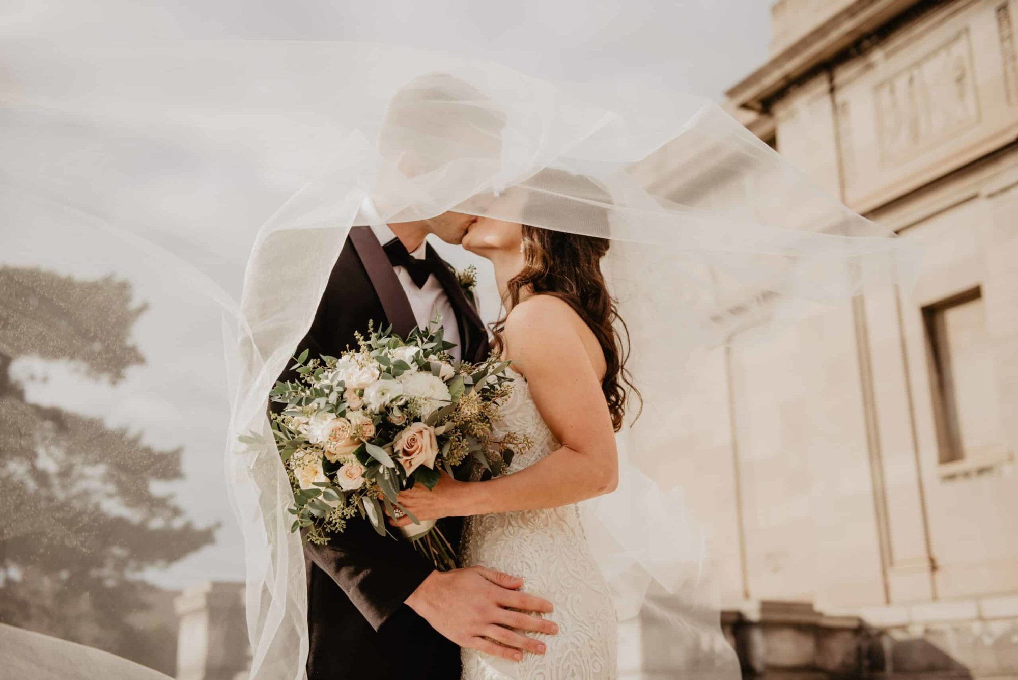 Bride and groom share a kiss under a flowing veil while holding a lush bouquet outside a stone building.