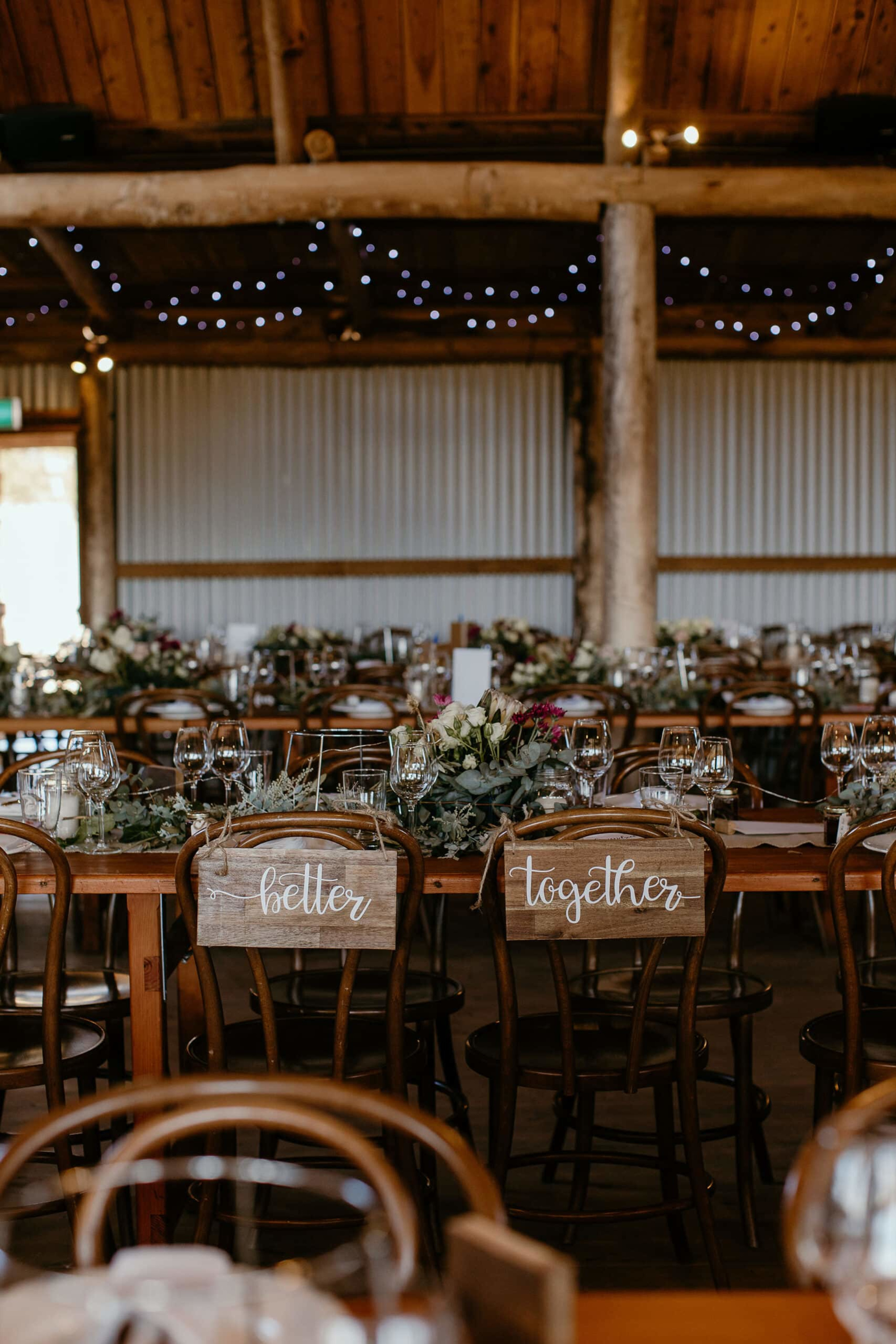 Rustic barn wedding reception with greenery-filled tables and wooden chairs marked with Better Together signs.