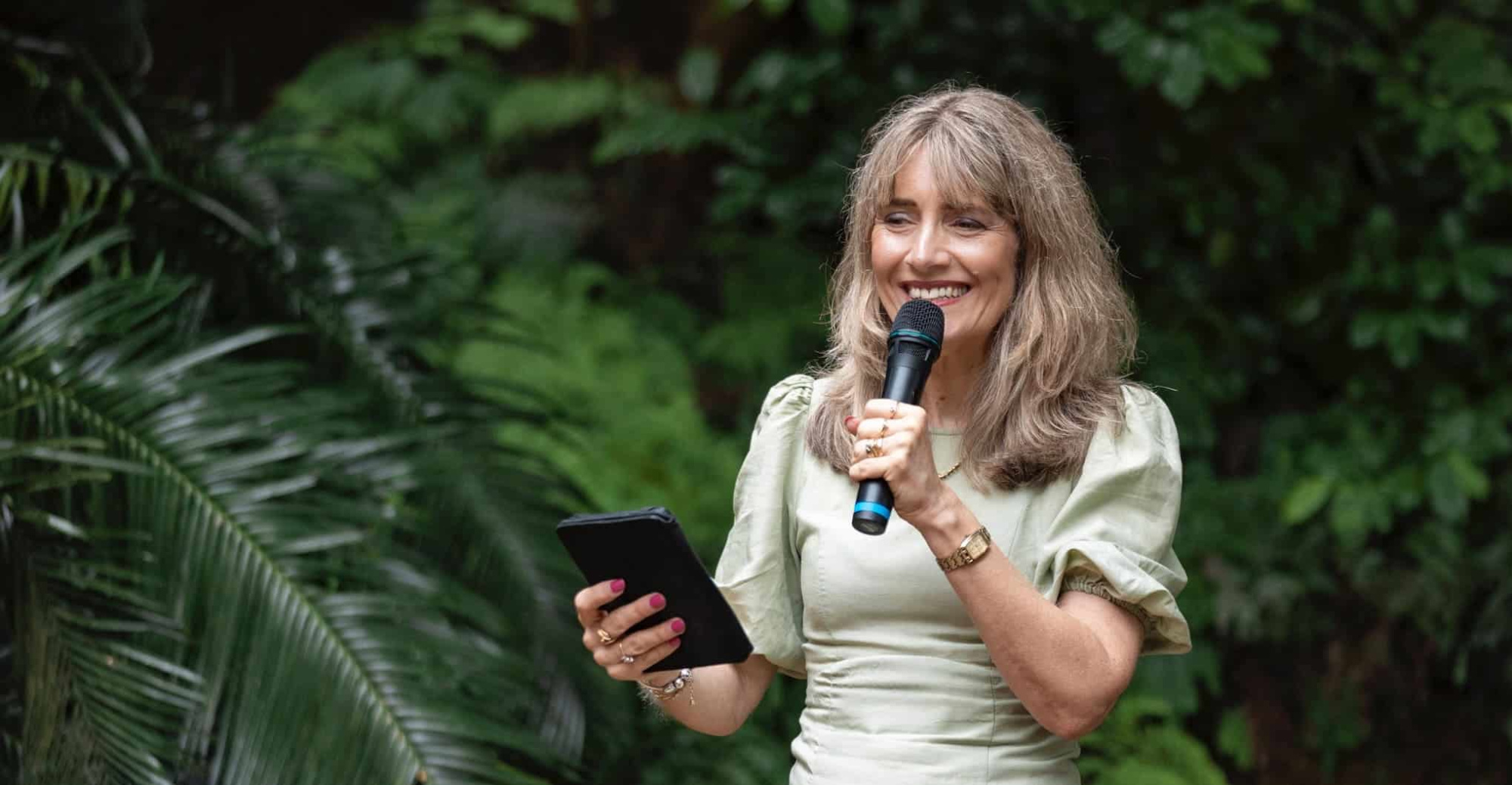 Smiling wedding celebrant holding a microphone and tablet during an outdoor ceremony in a lush garden setting.