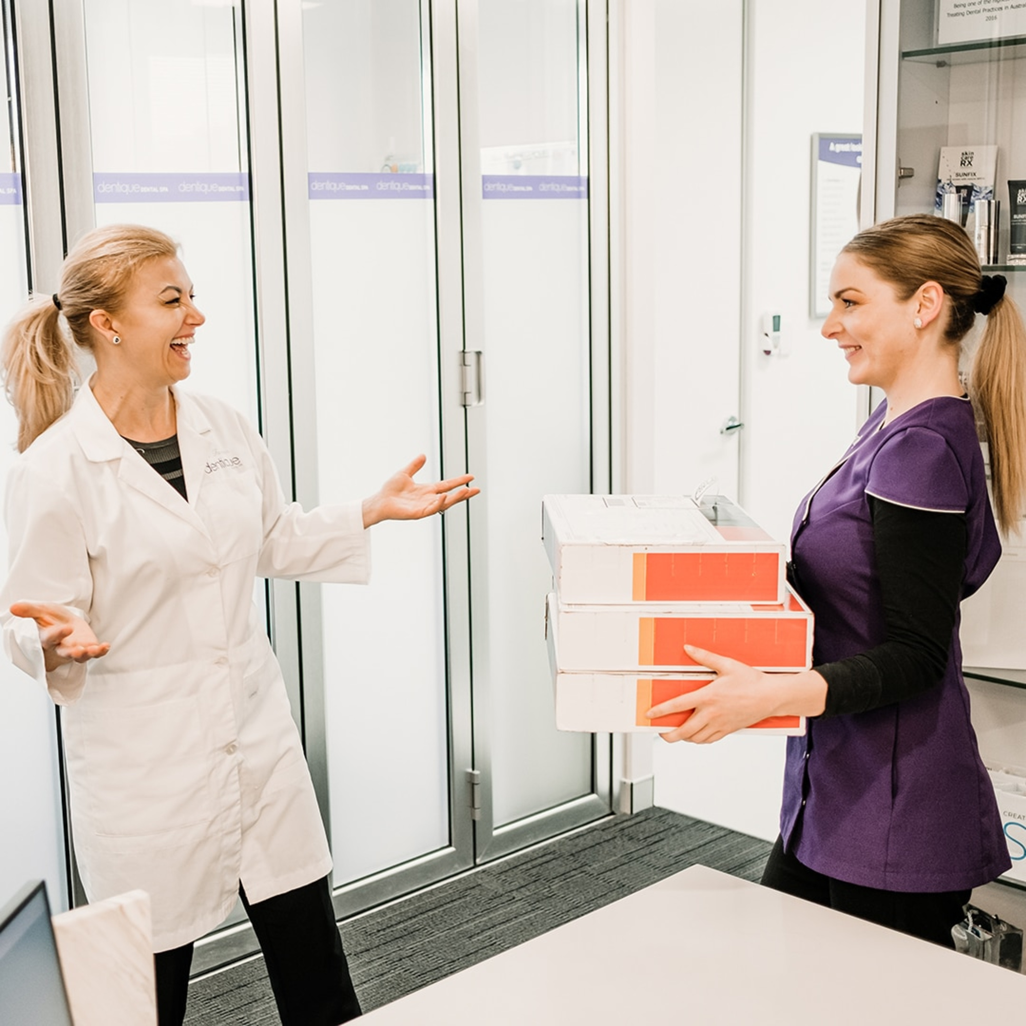 Two women in a modern office share a friendly conversation while one hands over a stack of boxes.
