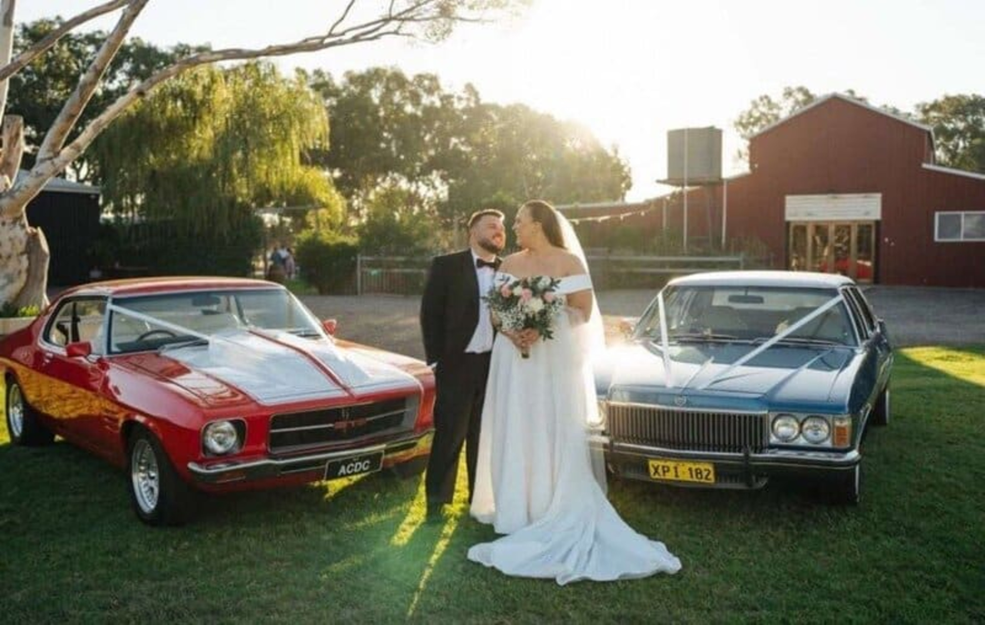 Bride and groom pose between two classic cars on the lawn outside a red barn at sunset.
