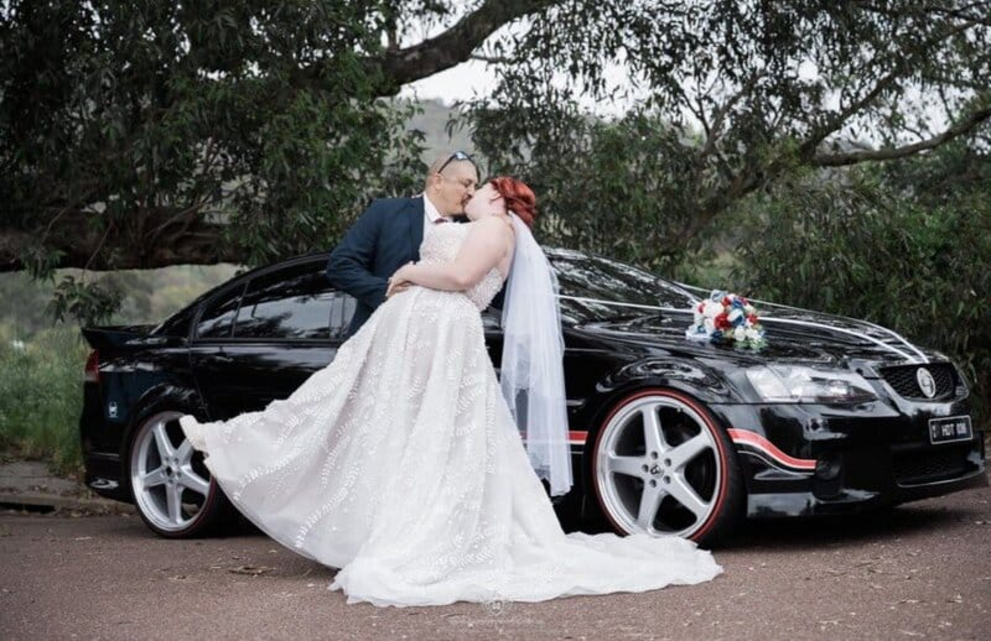 Bride and groom share a kiss while posing against a sleek black wedding car in an outdoor setting.