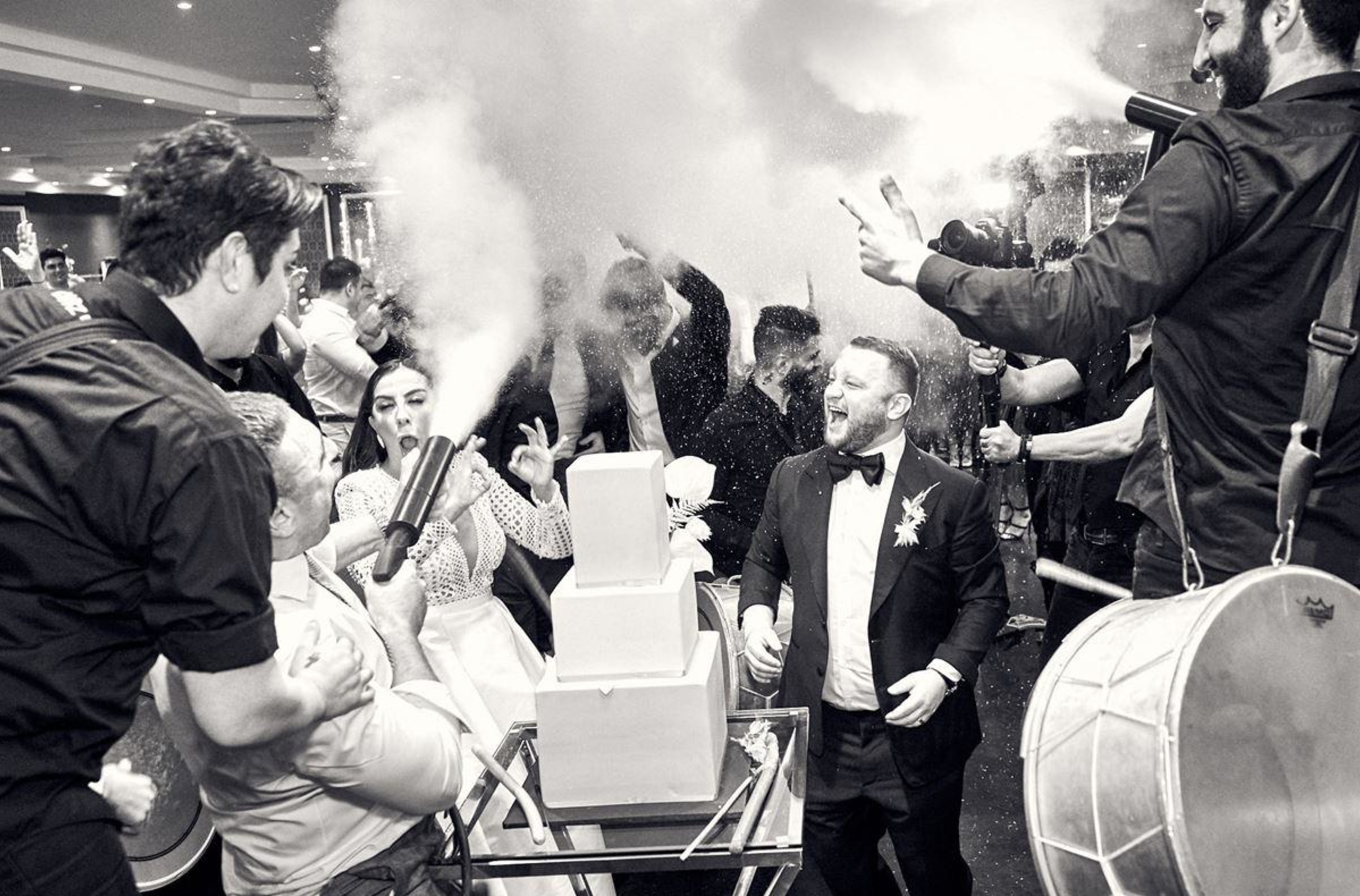 Candid black and white photo of a lively wedding reception with guests spraying smoke around the couple and cake.