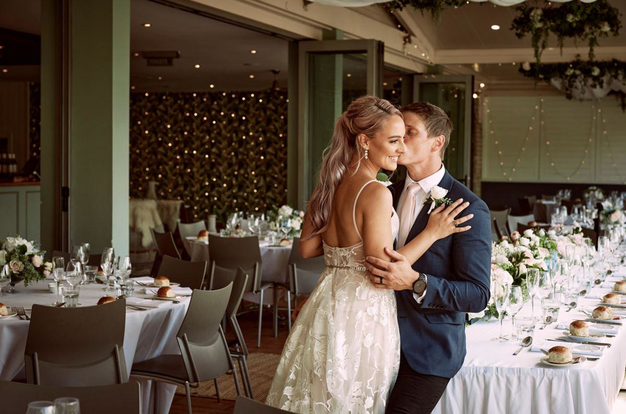 Bride and groom embrace in an elegantly decorated indoor wedding reception with long banquet tables and twinkling string lights.