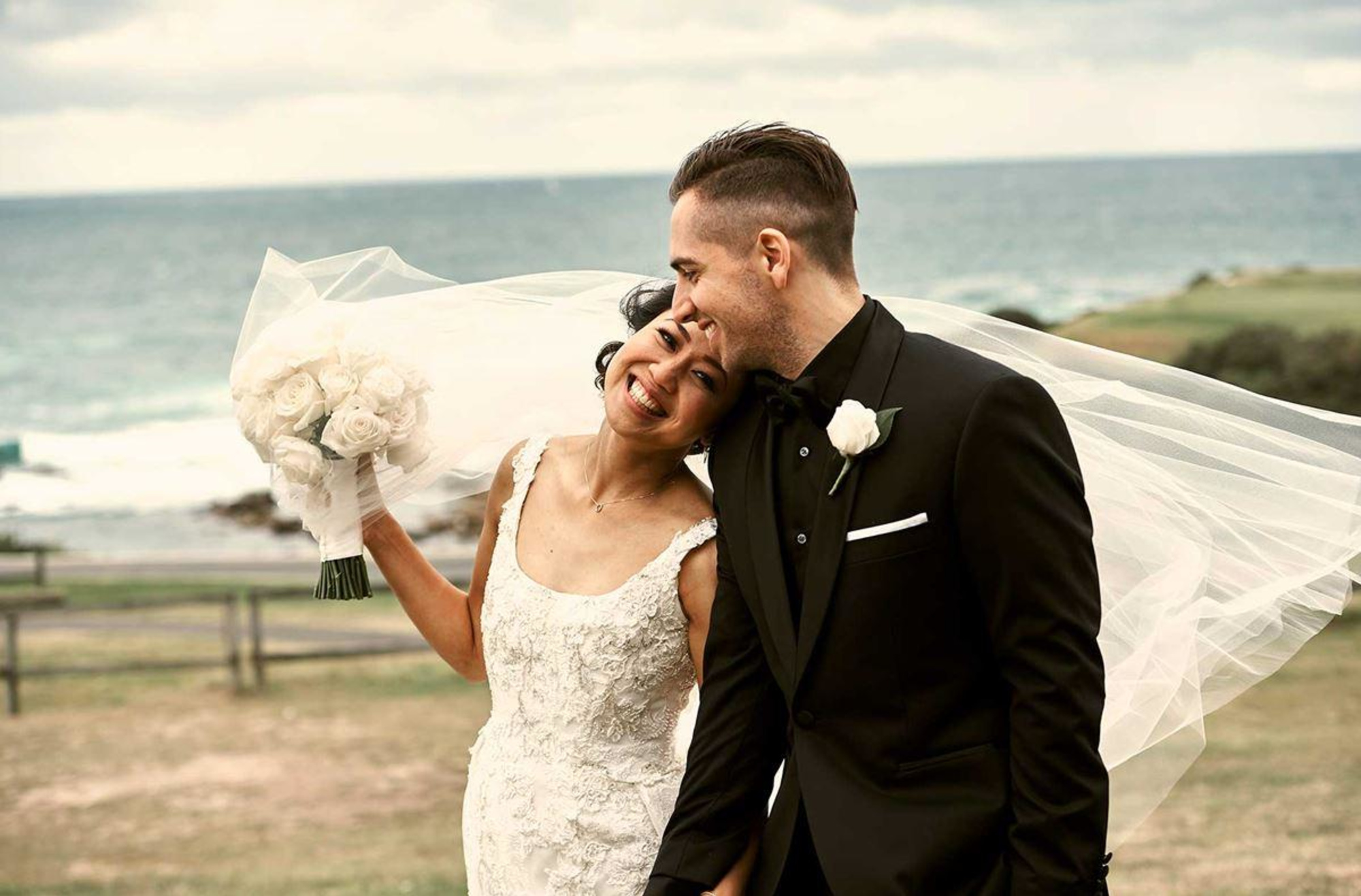 Smiling bride and groom embrace by the seaside with her veil and bouquet blowing in the wind.