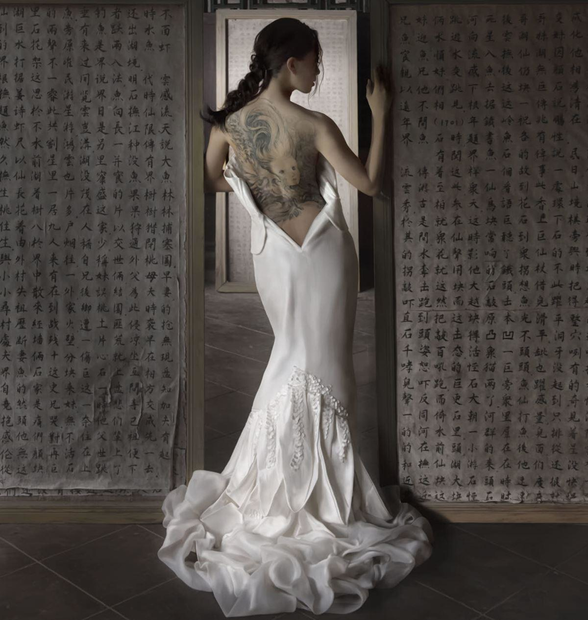 Tattooed bride in a backless white gown stands between panels of calligraphy in a dramatic artistic portrait.