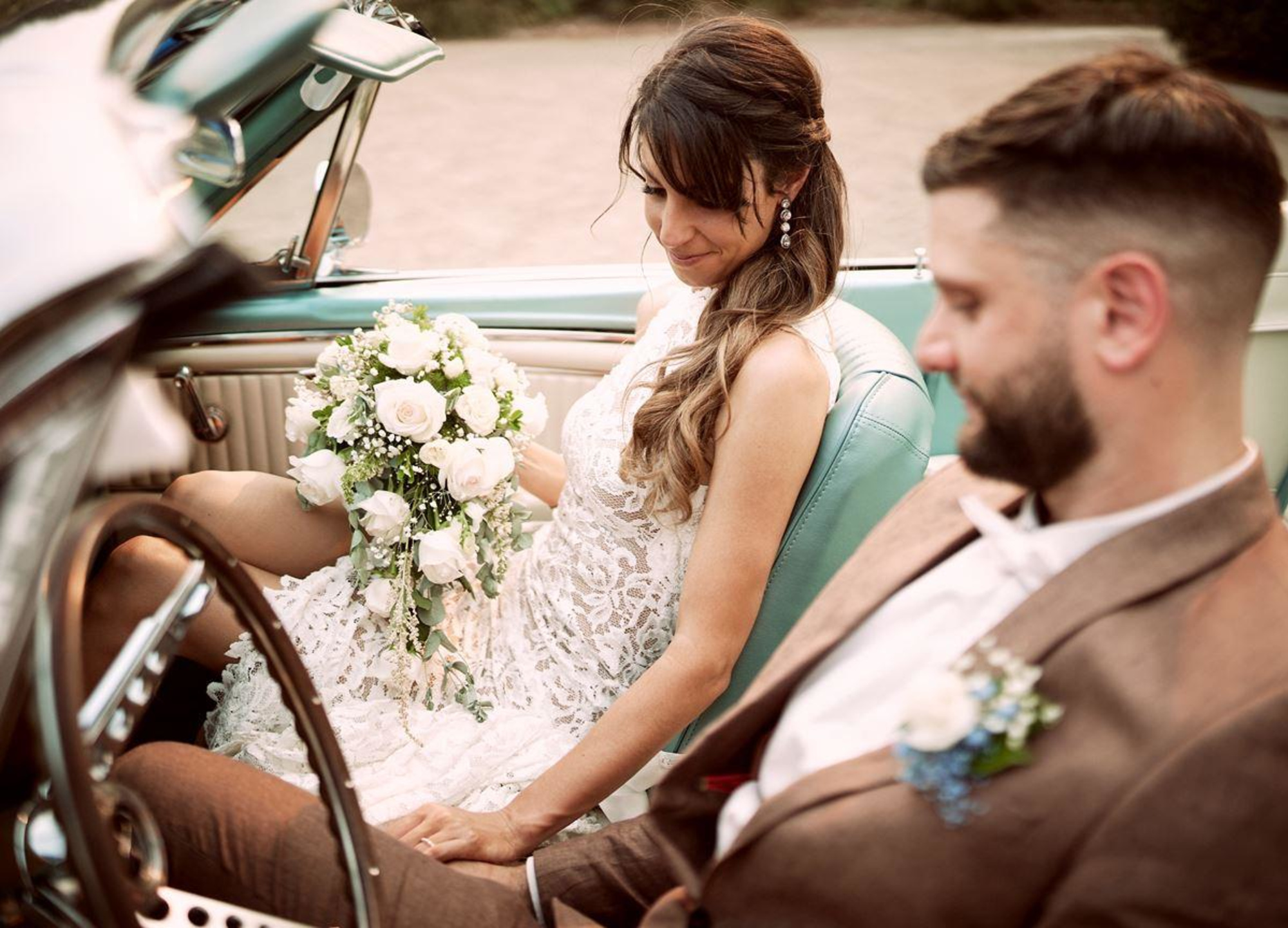 Bride holding a cascading white bouquet while sitting with her partner in a vintage convertible.