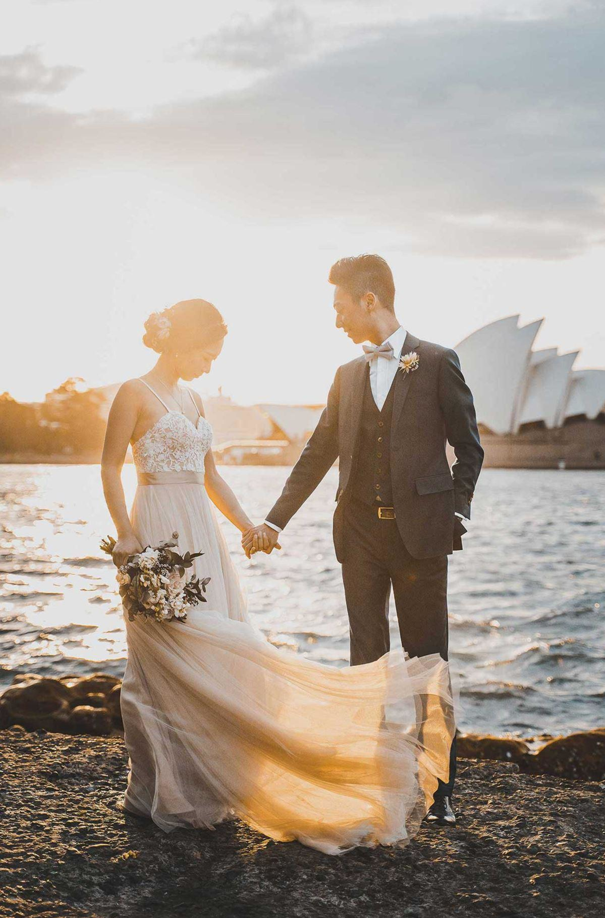 Bride and groom hold hands by the water at sunset with her flowing dress lit by golden light.