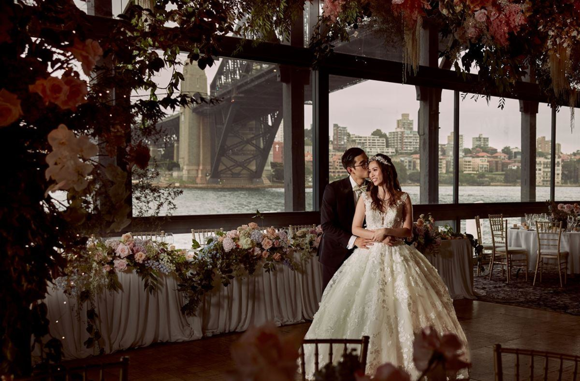 Bride and groom embrace in a waterfront reception venue with lush florals and city views through large windows.