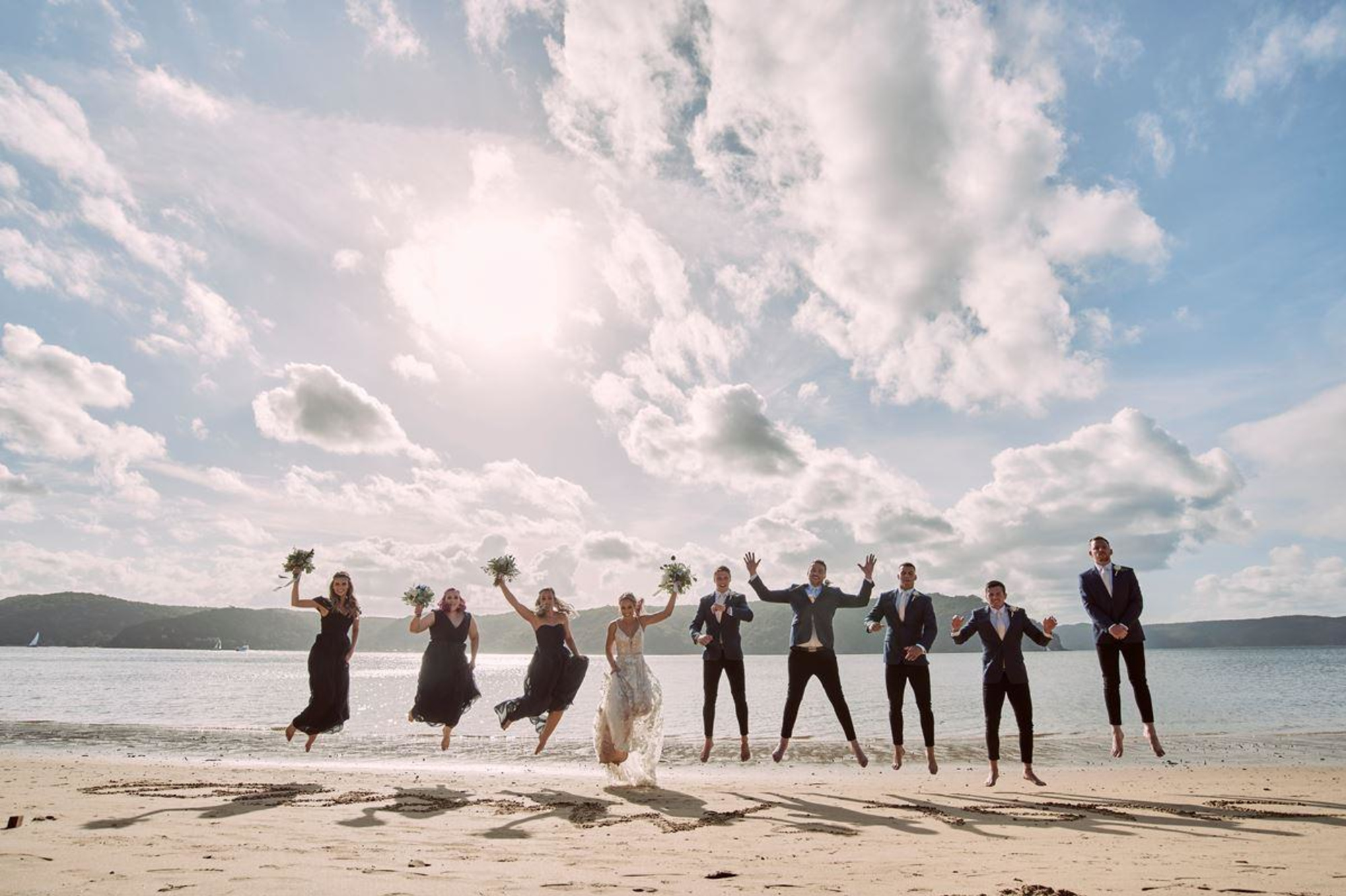 Wedding party jumps together on a sunny beach shoreline with ocean and clouds in the background.
