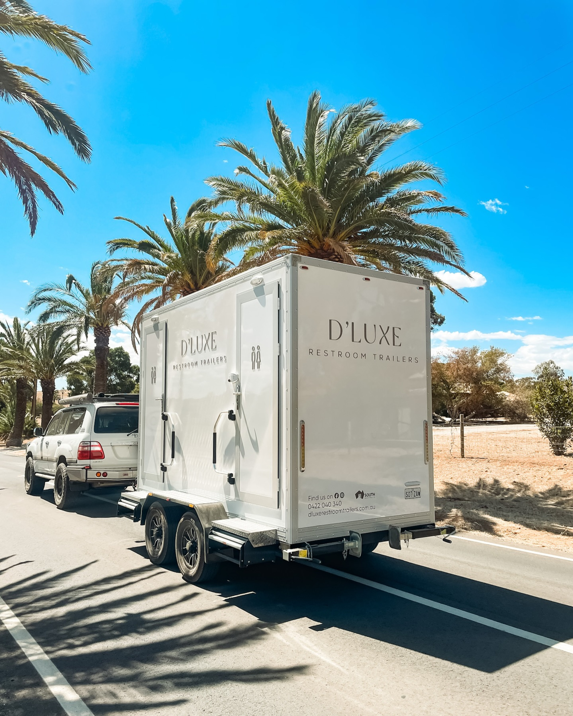 Luxury D'Luxe restroom trailer being towed along a palm-lined road for an outdoor event.