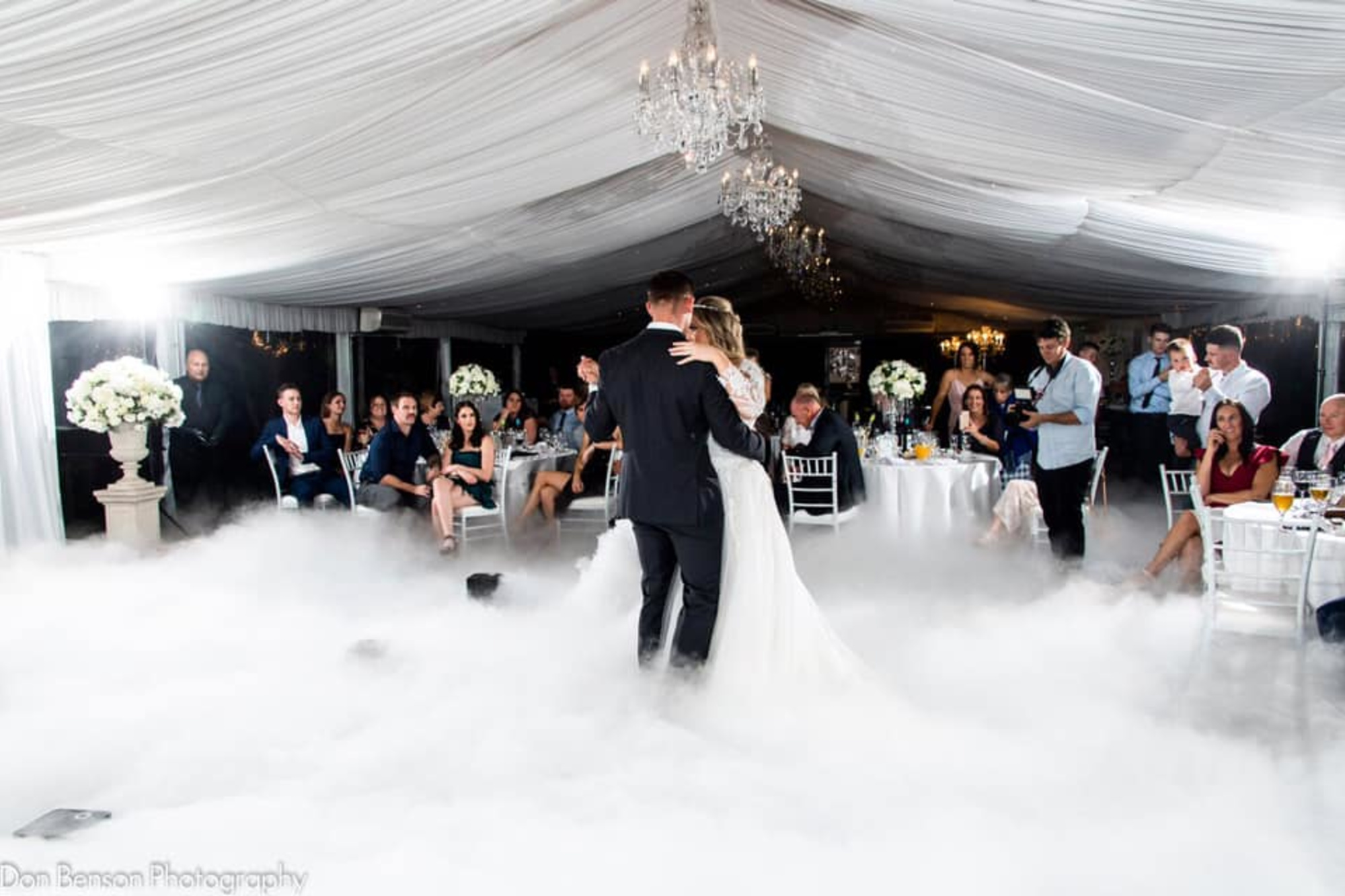 Bride and groom share their first dance on a fog-covered dance floor in an elegant tented reception surrounded by guests.