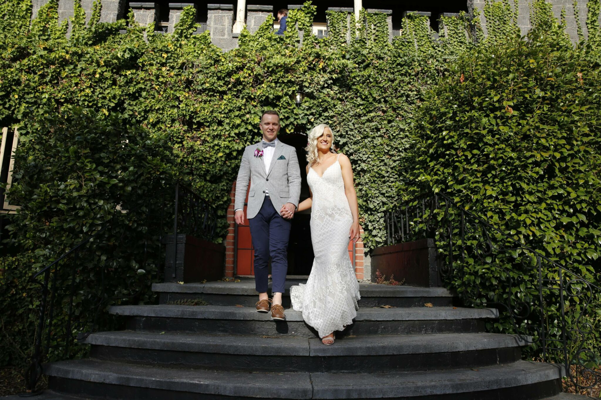 Bride and groom walk down stone steps in front of an ivy-covered building at their outdoor wedding.
