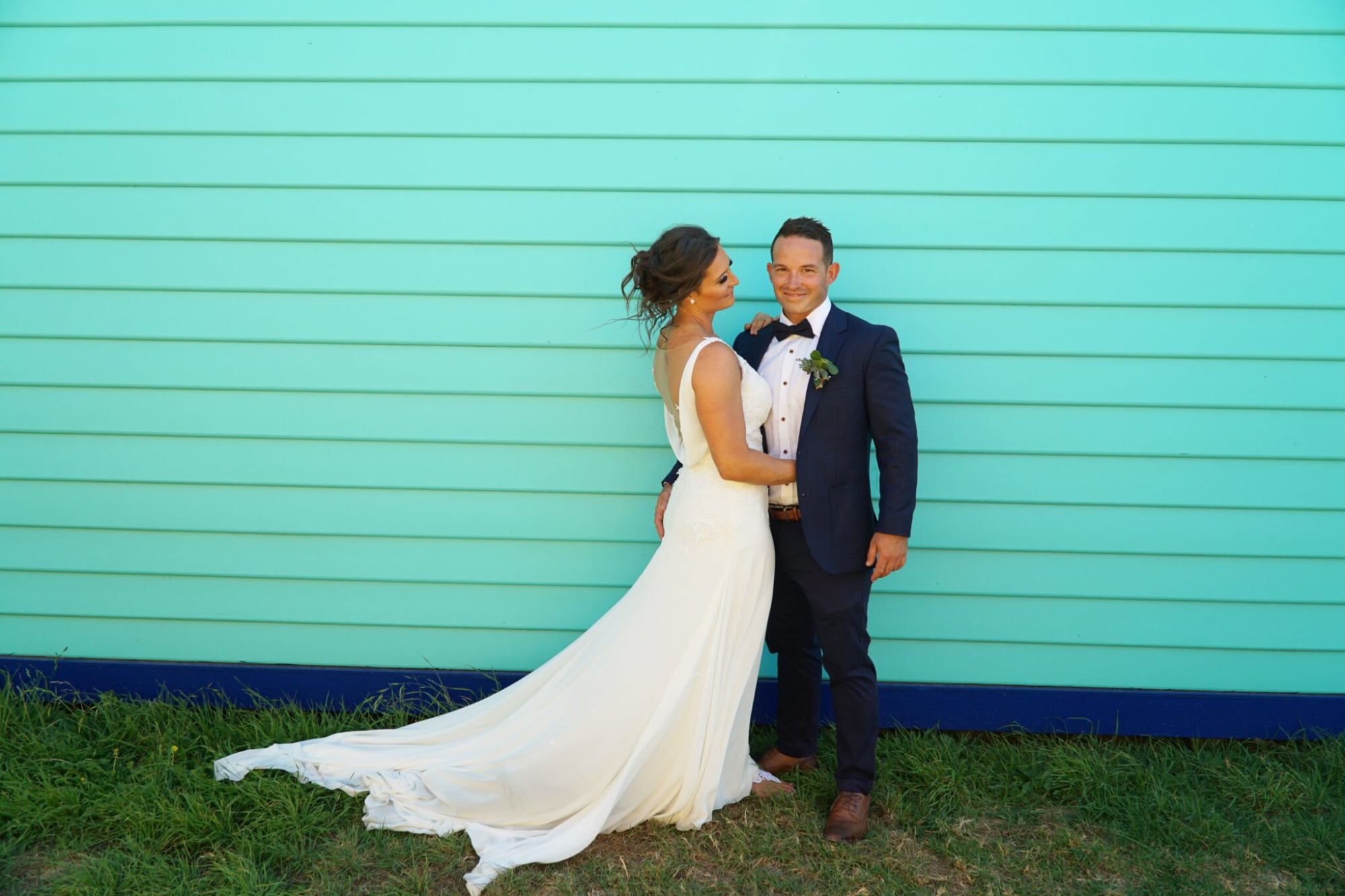 Bride and groom posing together against a bright turquoise wooden wall outdoors.