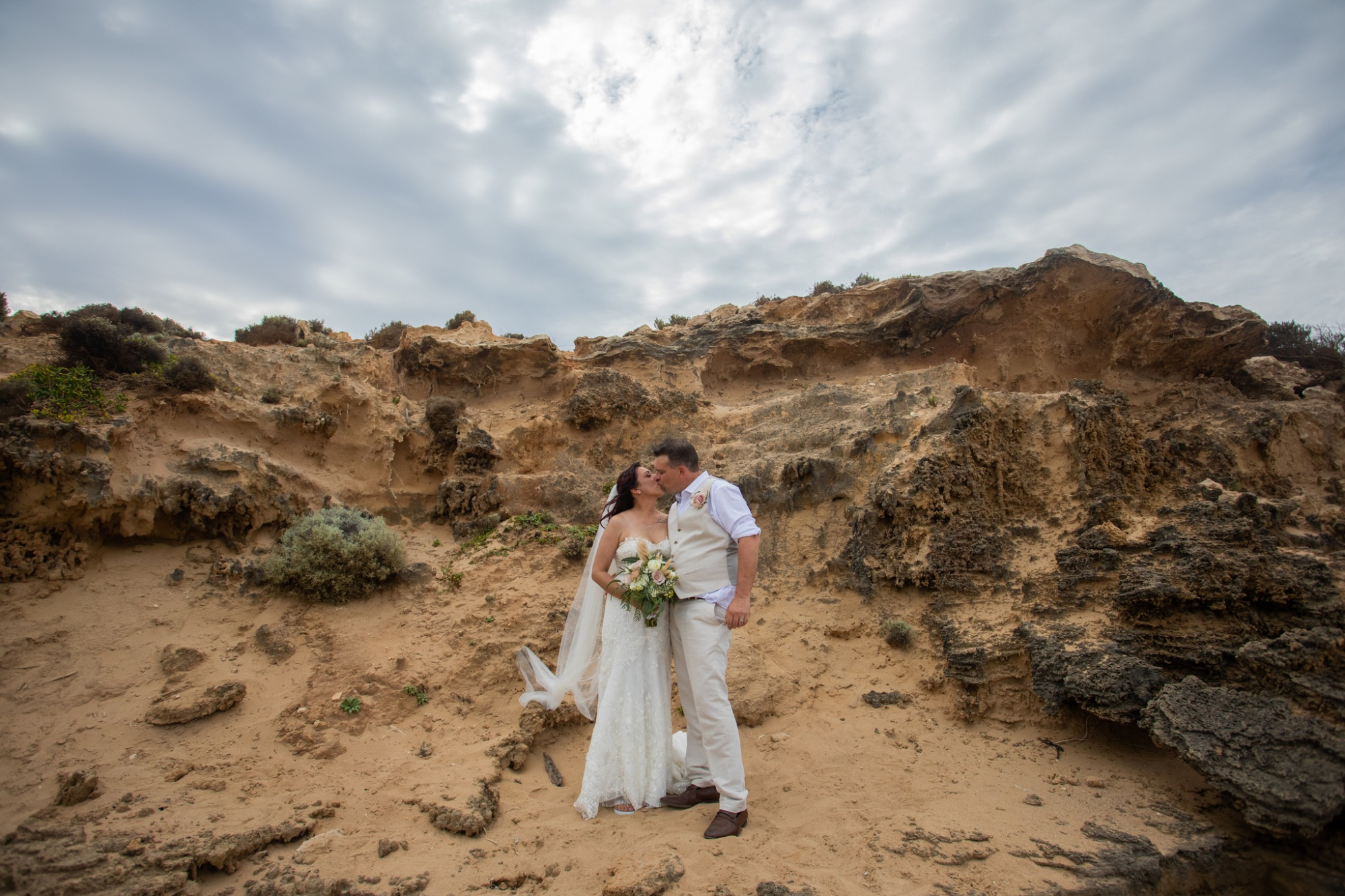 Bride and groom share a kiss against rugged coastal cliffs and a cloudy sky.