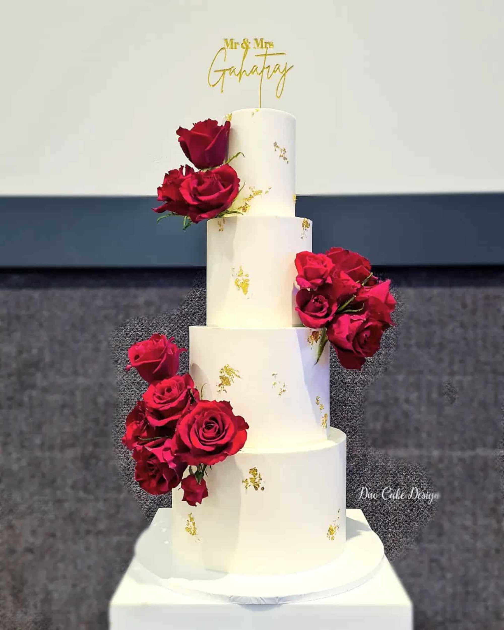Four-tier white wedding cake with red roses and gold leaf accents topped with a custom Mr and Mrs cake topper.