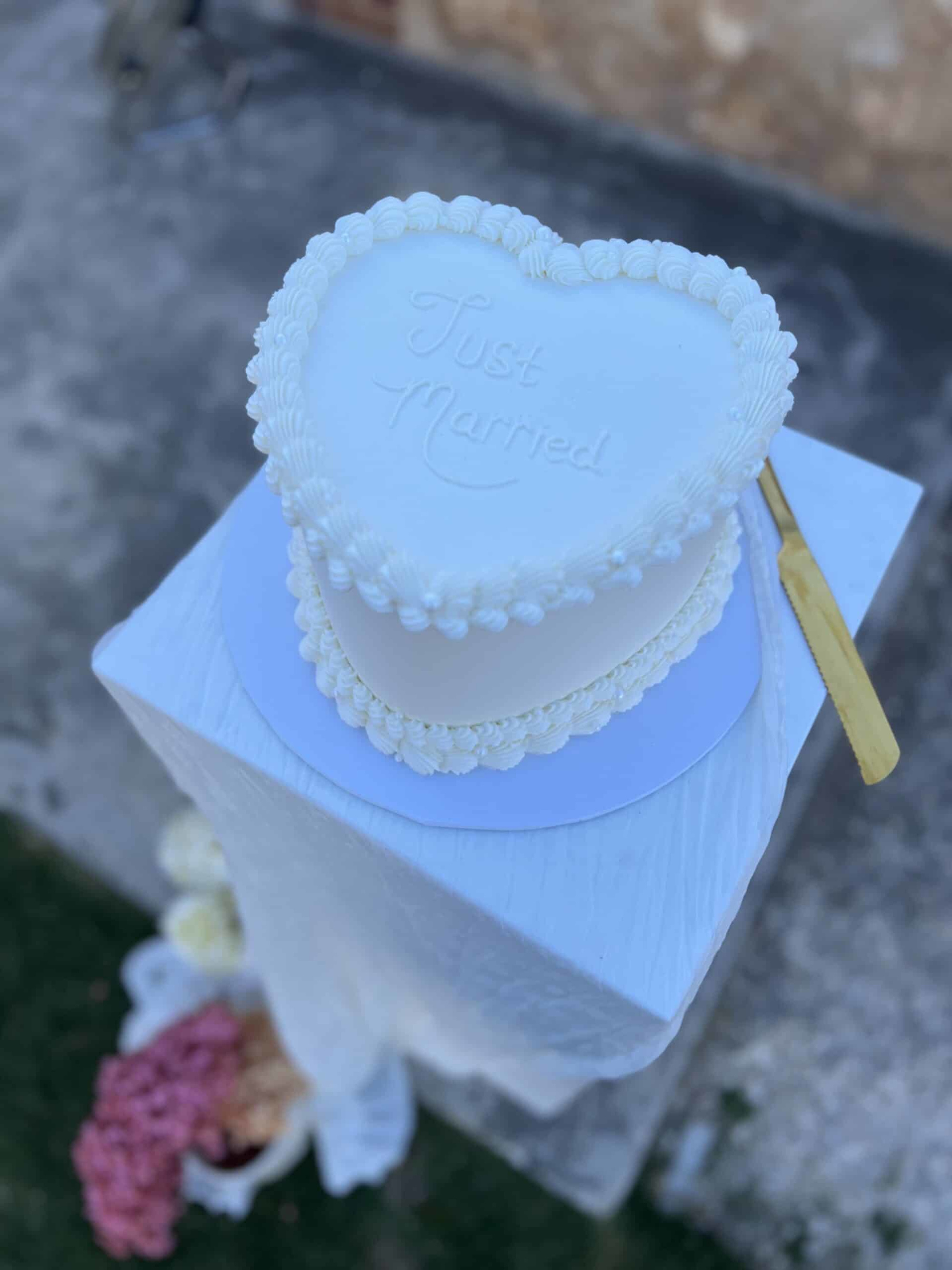 Heart-shaped white wedding cake with 'Just Married' icing displayed on a tall pedestal outdoors.