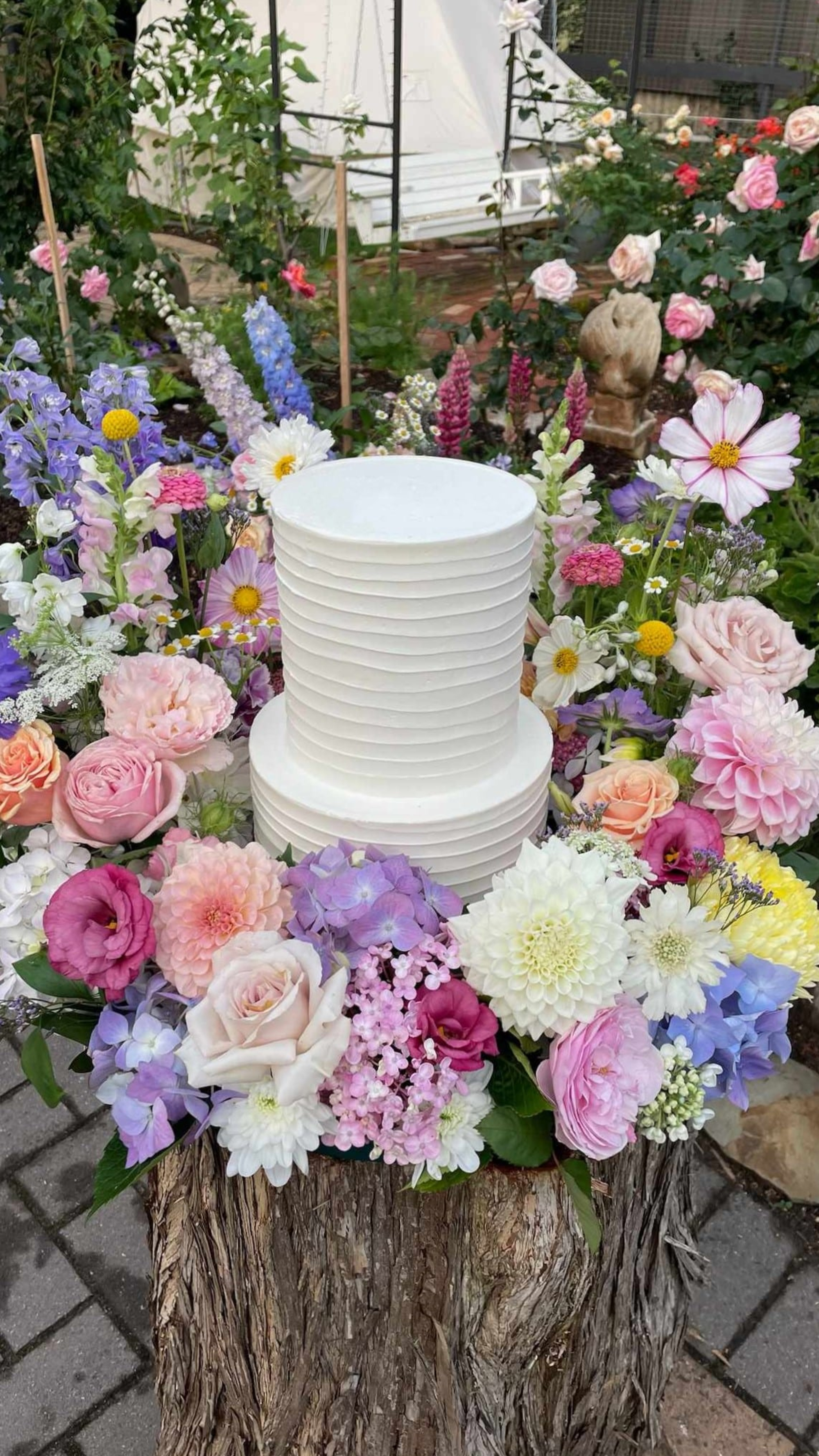 Two-tier white buttercream wedding cake surrounded by lush pastel flowers in a garden setting.