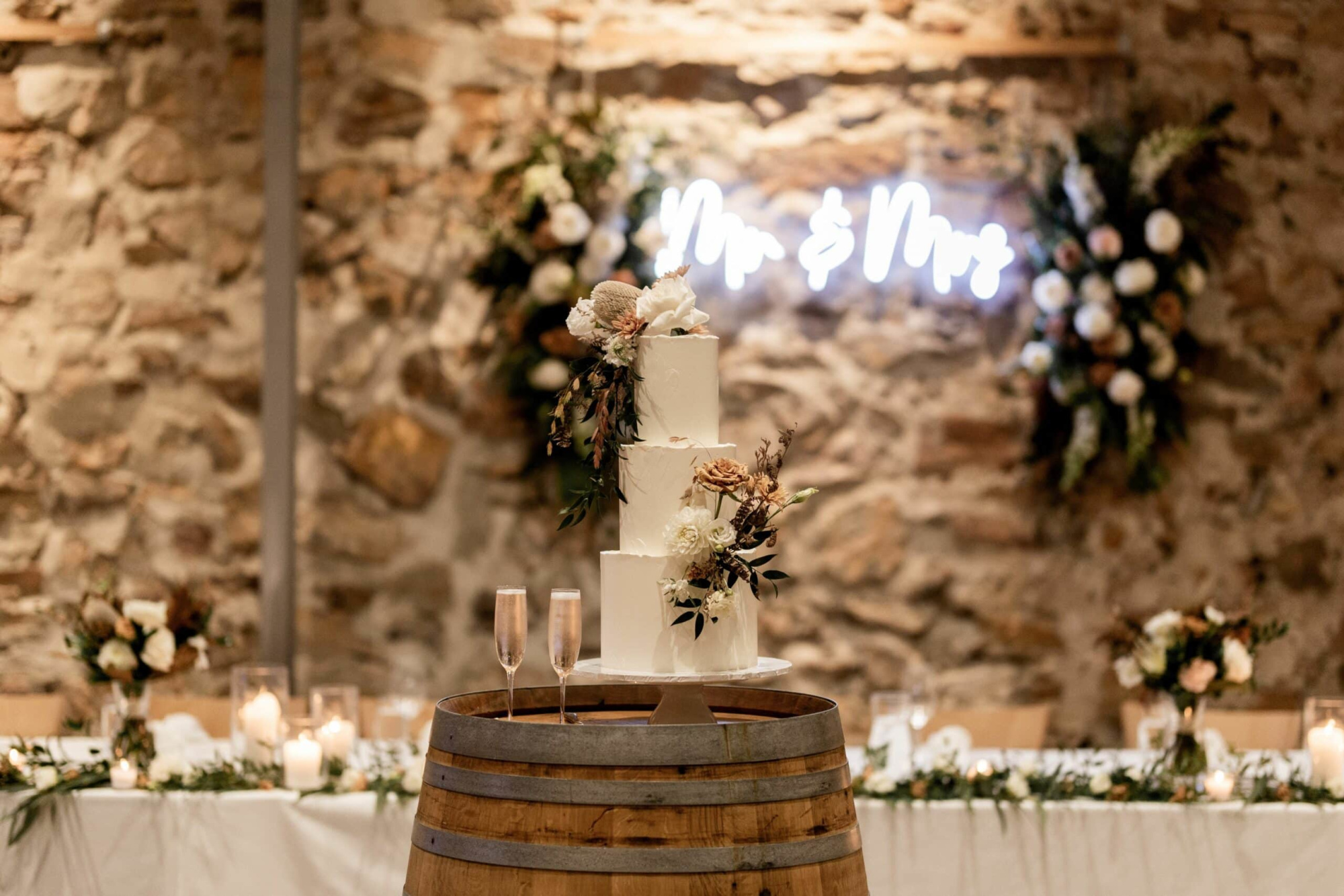 Three-tier white wedding cake with florals on a barrel in a rustic stone reception with candles and greenery.