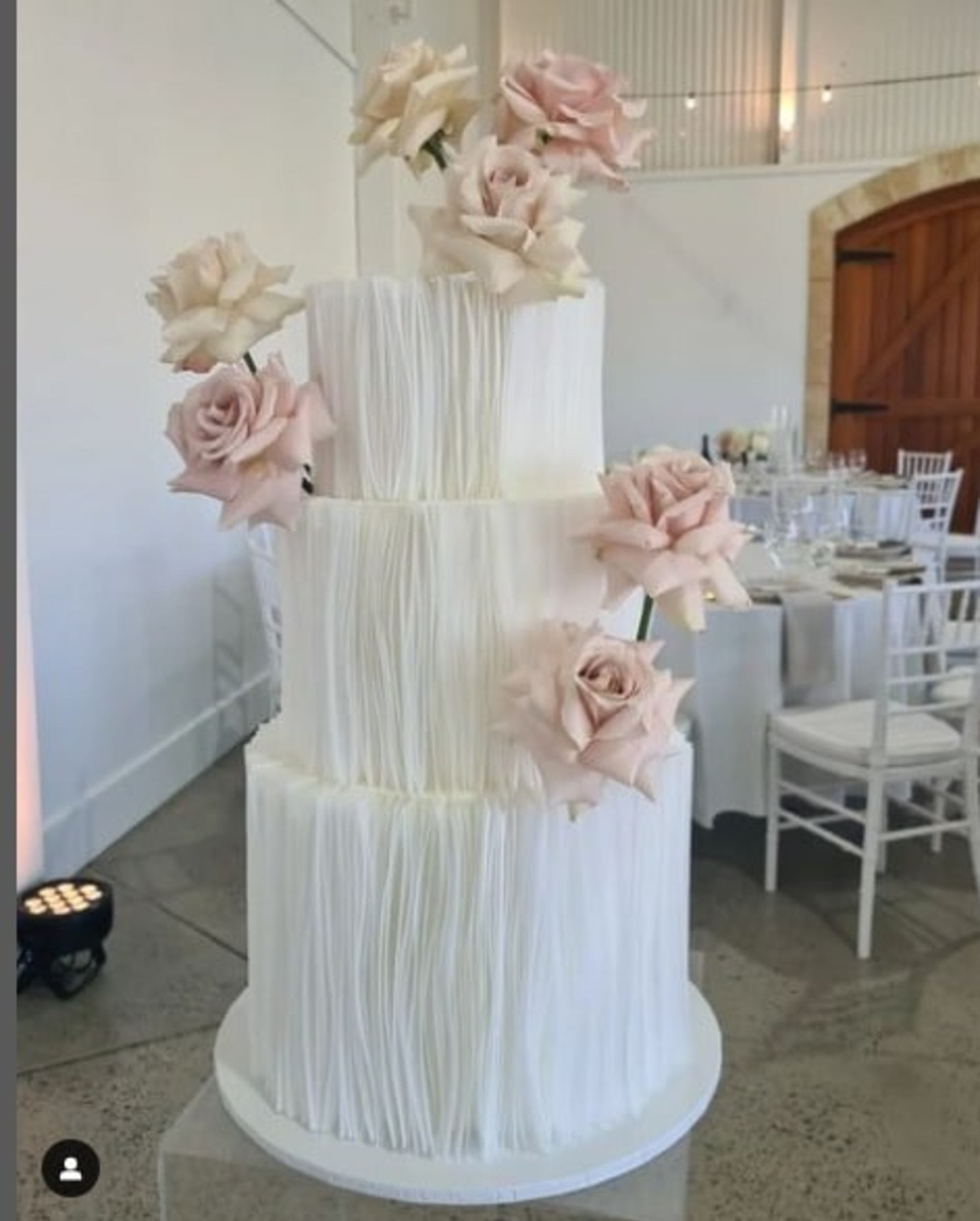 Three-tier white wedding cake with textured vertical detailing and blush rose accents in a modern reception space.