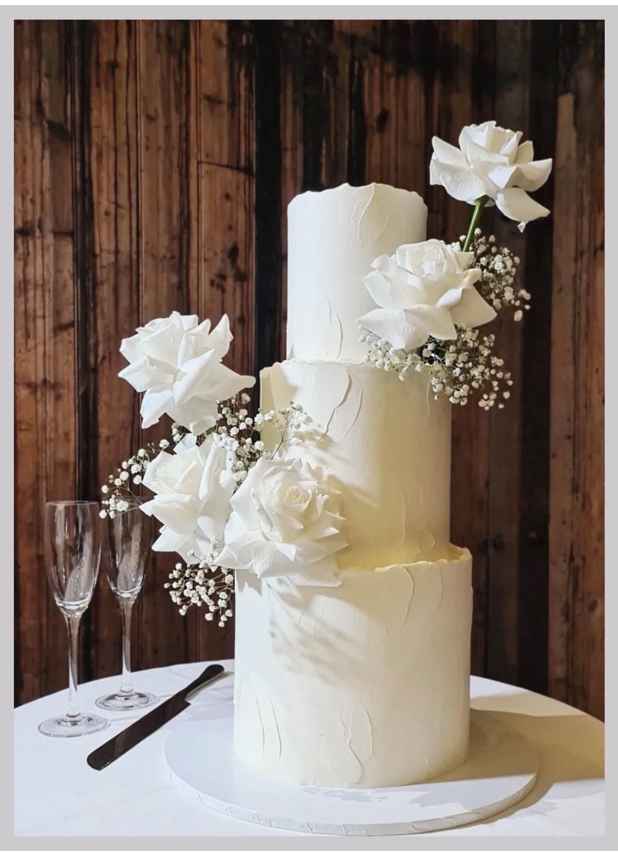 Tall three-tier white wedding cake with white roses and baby's breath on a table in front of a rustic wooden backdrop.