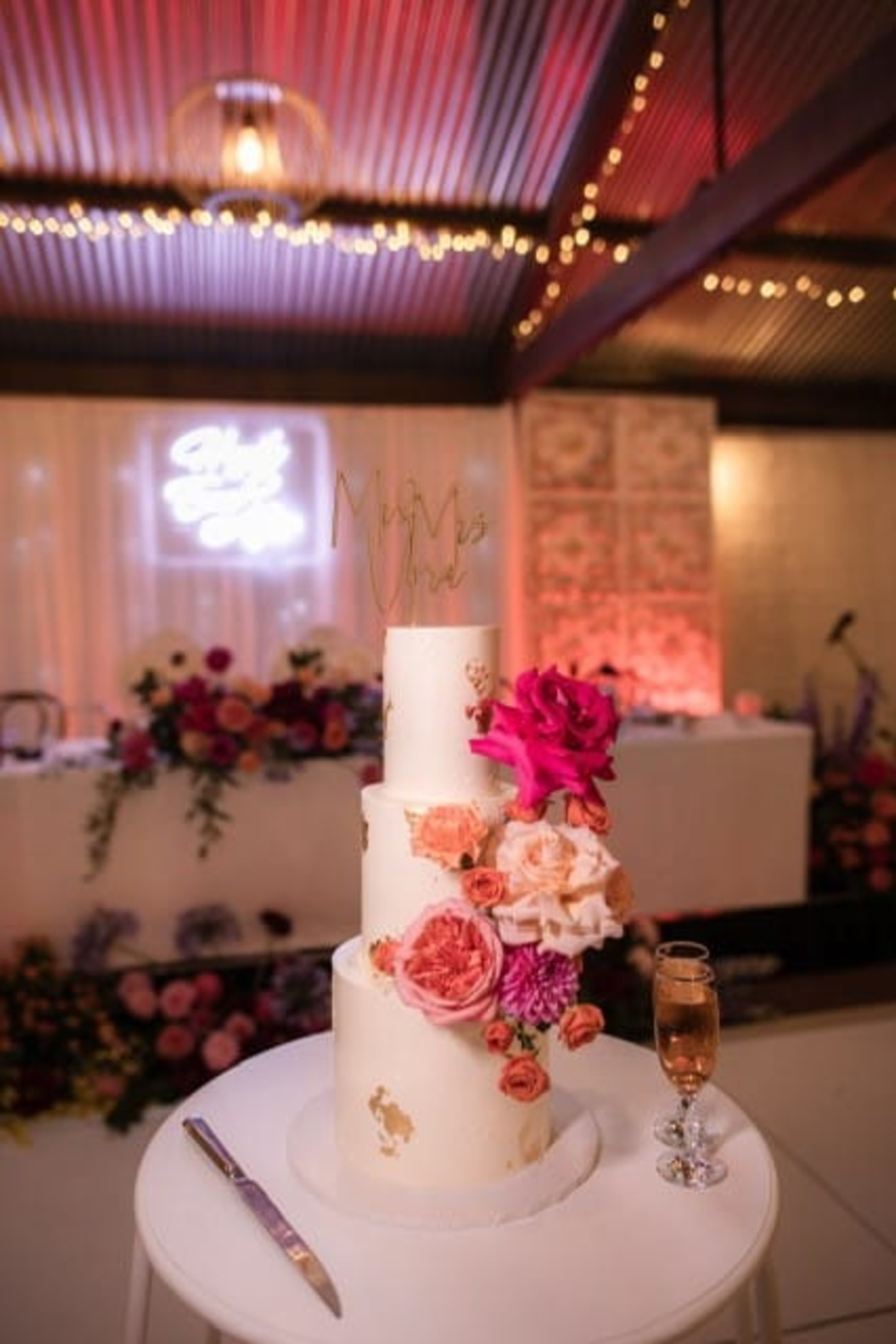 Three-tier white wedding cake with colorful floral accents and gold topper on a table at a romantic indoor reception.