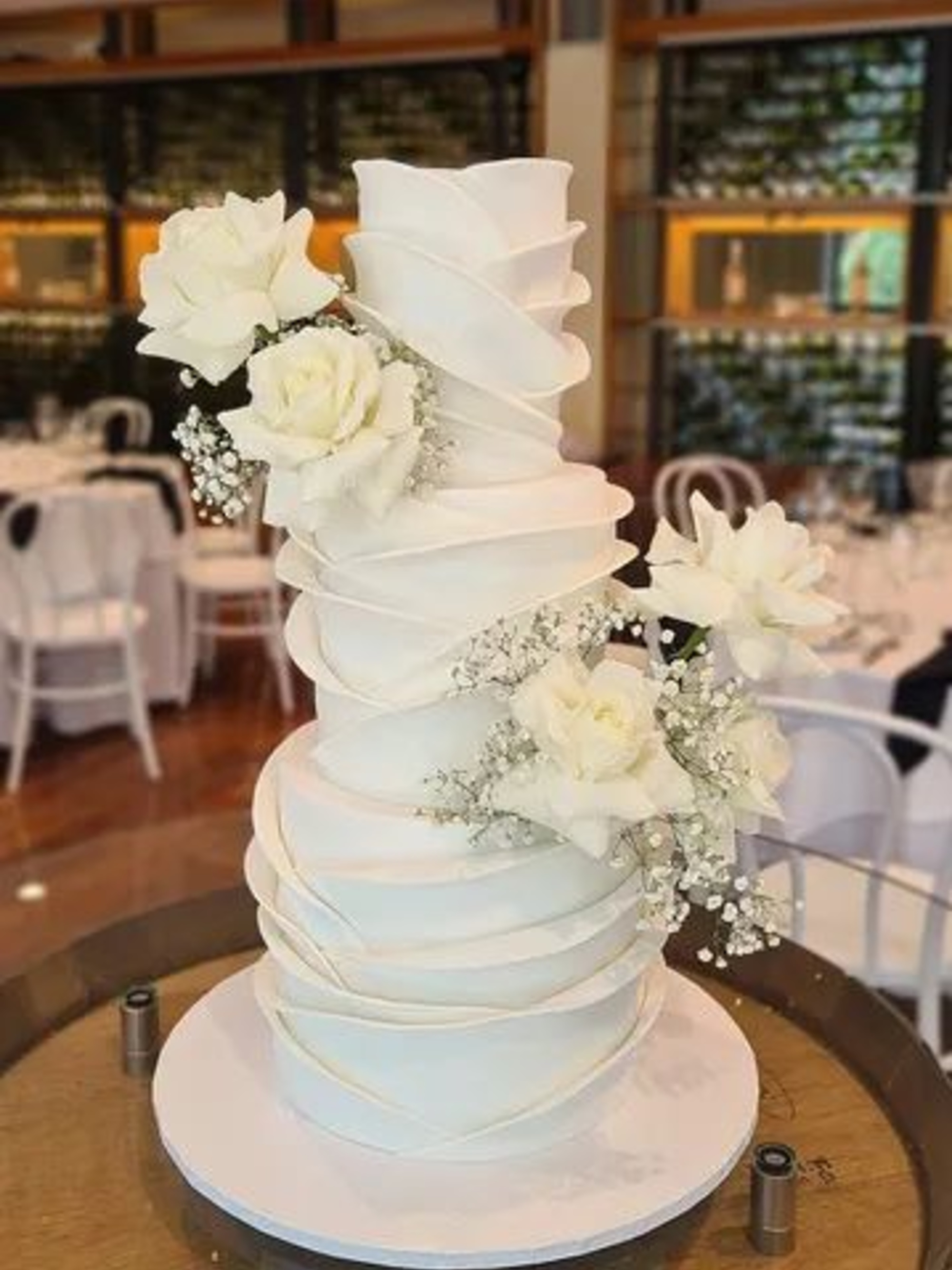 Tall modern white wedding cake with spiral fondant ribbons and white rose flowers at a reception venue.