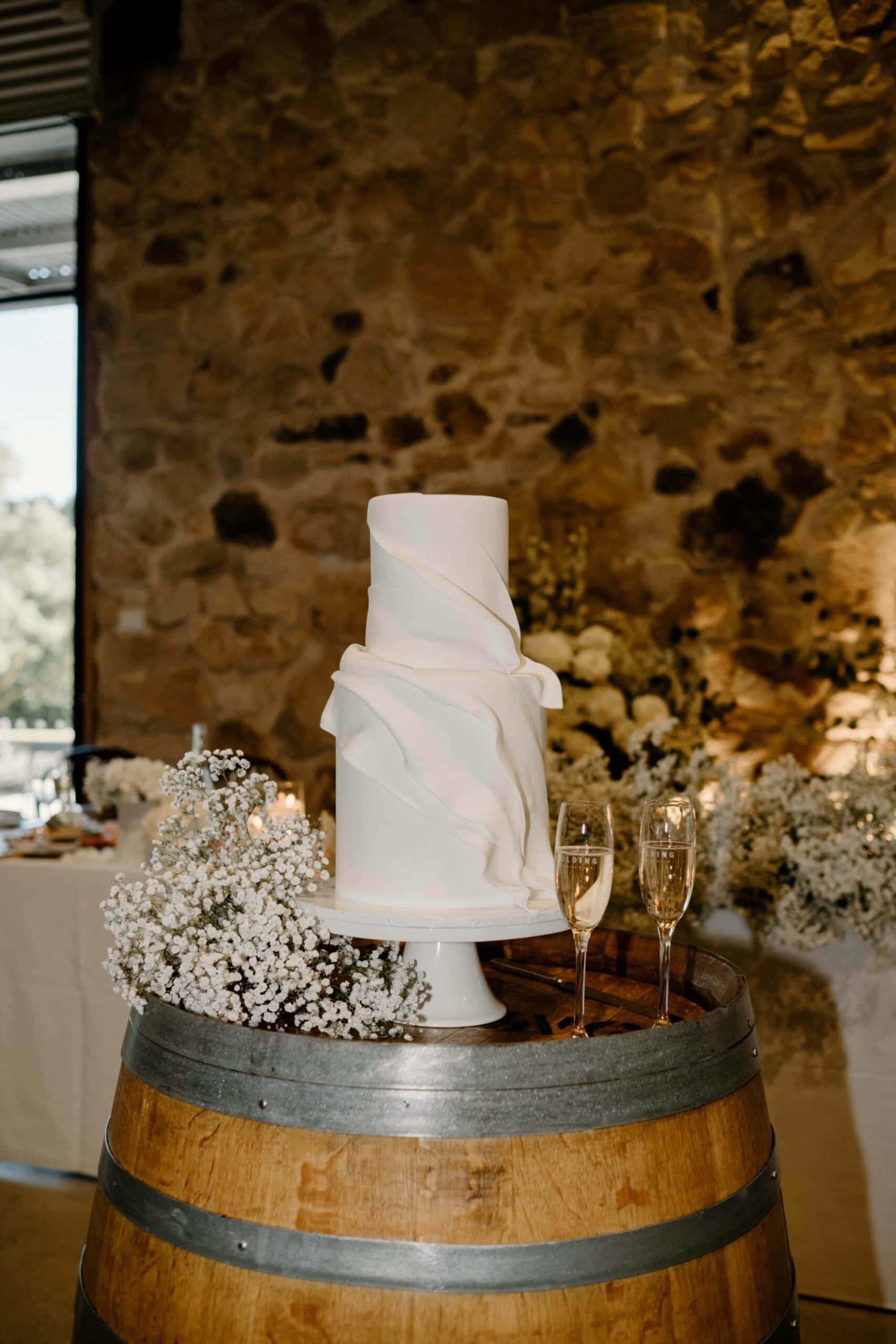 Tall white minimalist wedding cake on a wine barrel with baby’s breath and champagne flutes in a rustic stone venue.