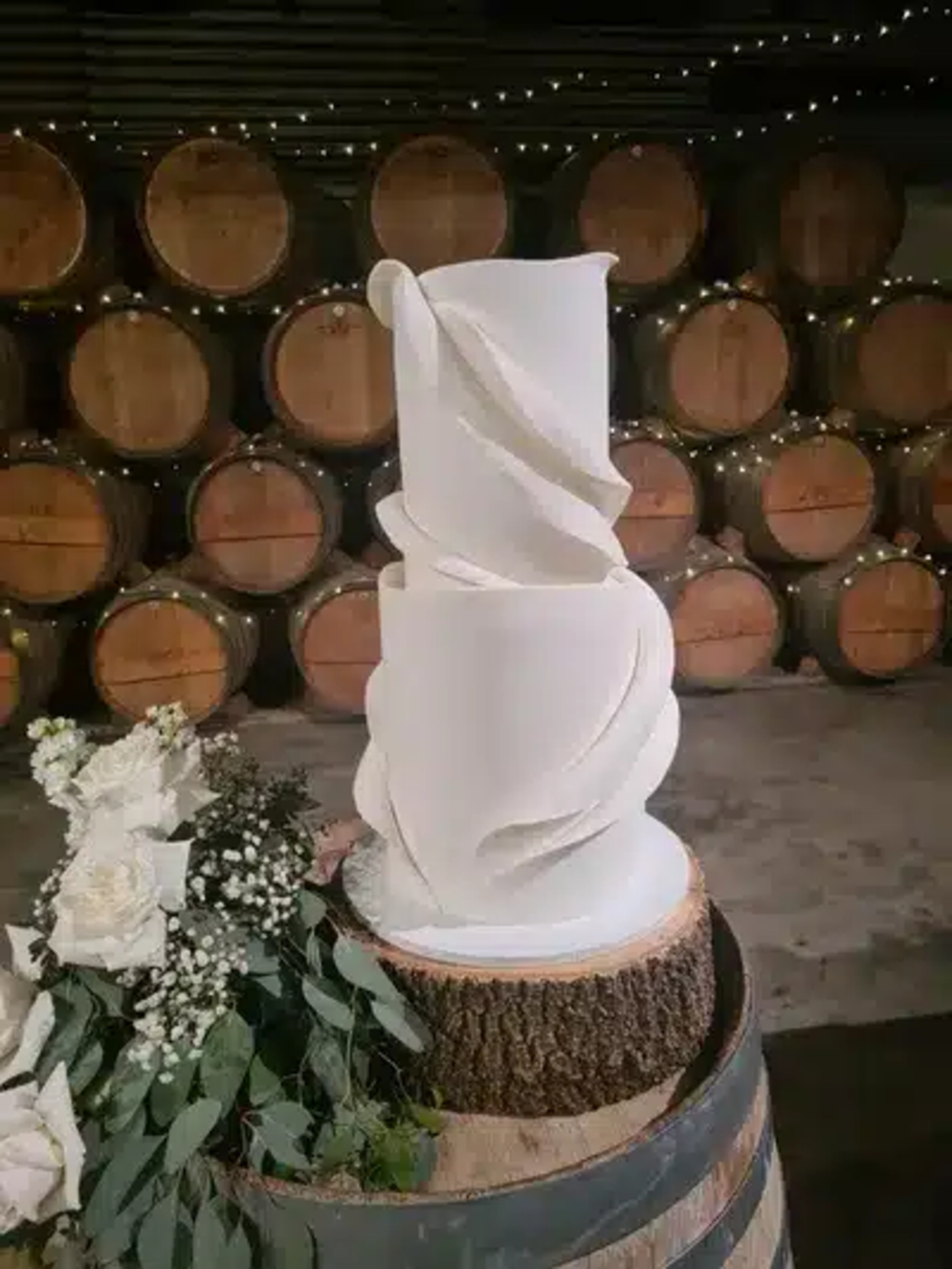 Modern white three-tier wedding cake with sculpted details in a barrel room decorated with flowers and string lights.