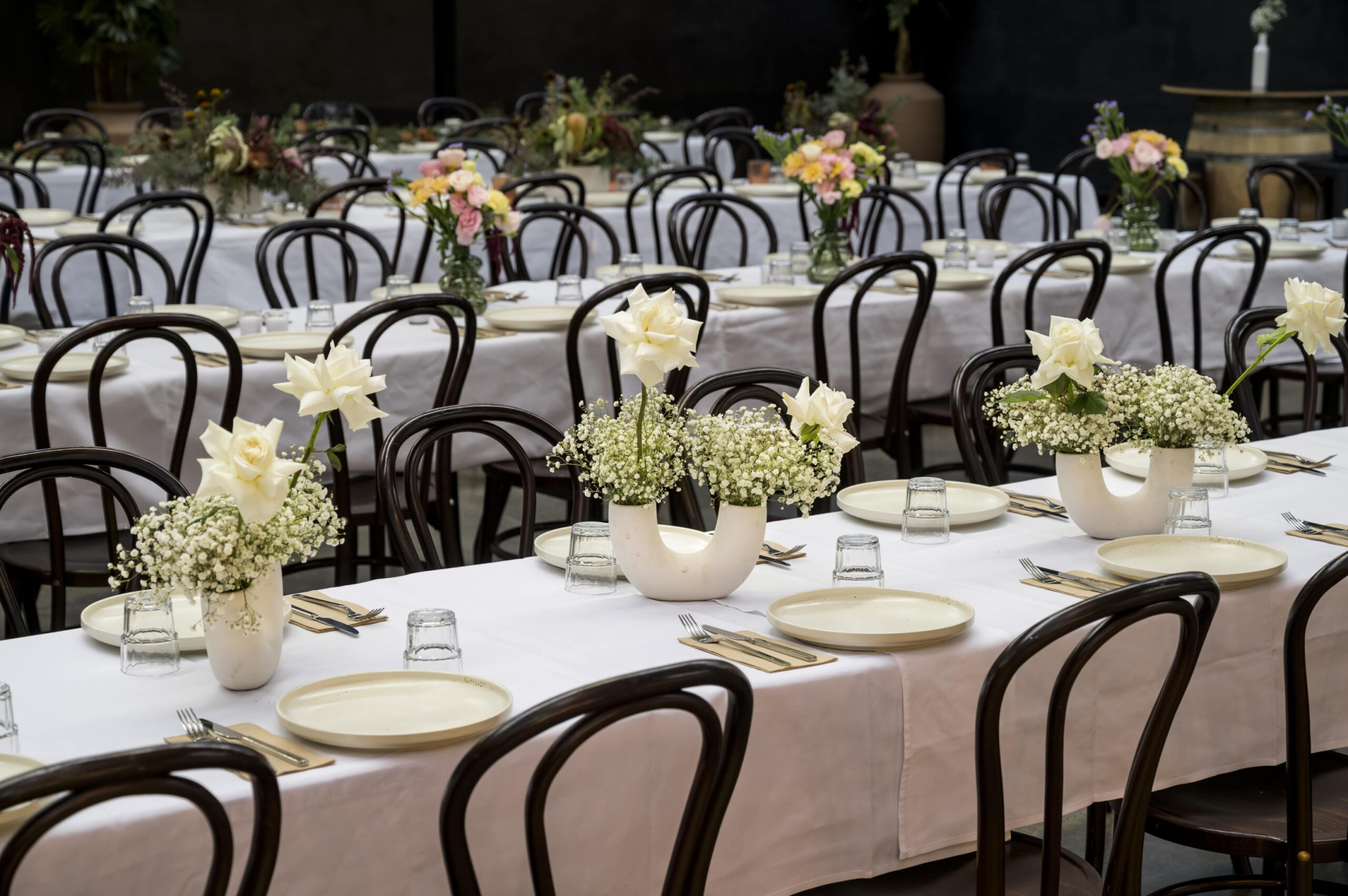 Elegant wedding reception tables with white linens, simple dishware, and white rose centerpieces in a spacious indoor venue.