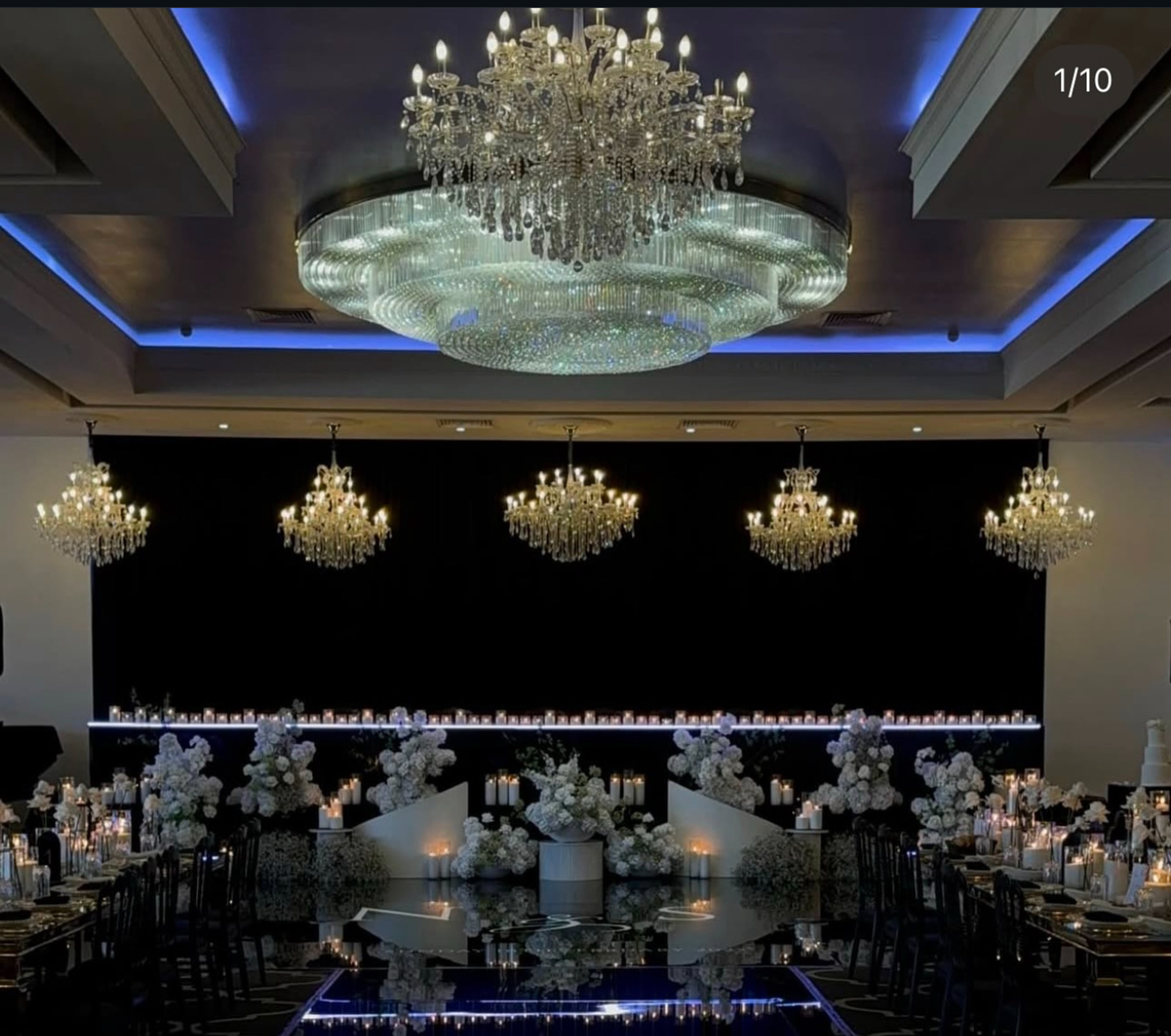 Luxurious indoor wedding reception with grand chandeliers, white floral arrangements, and candlelit tables around a reflective dance floor.