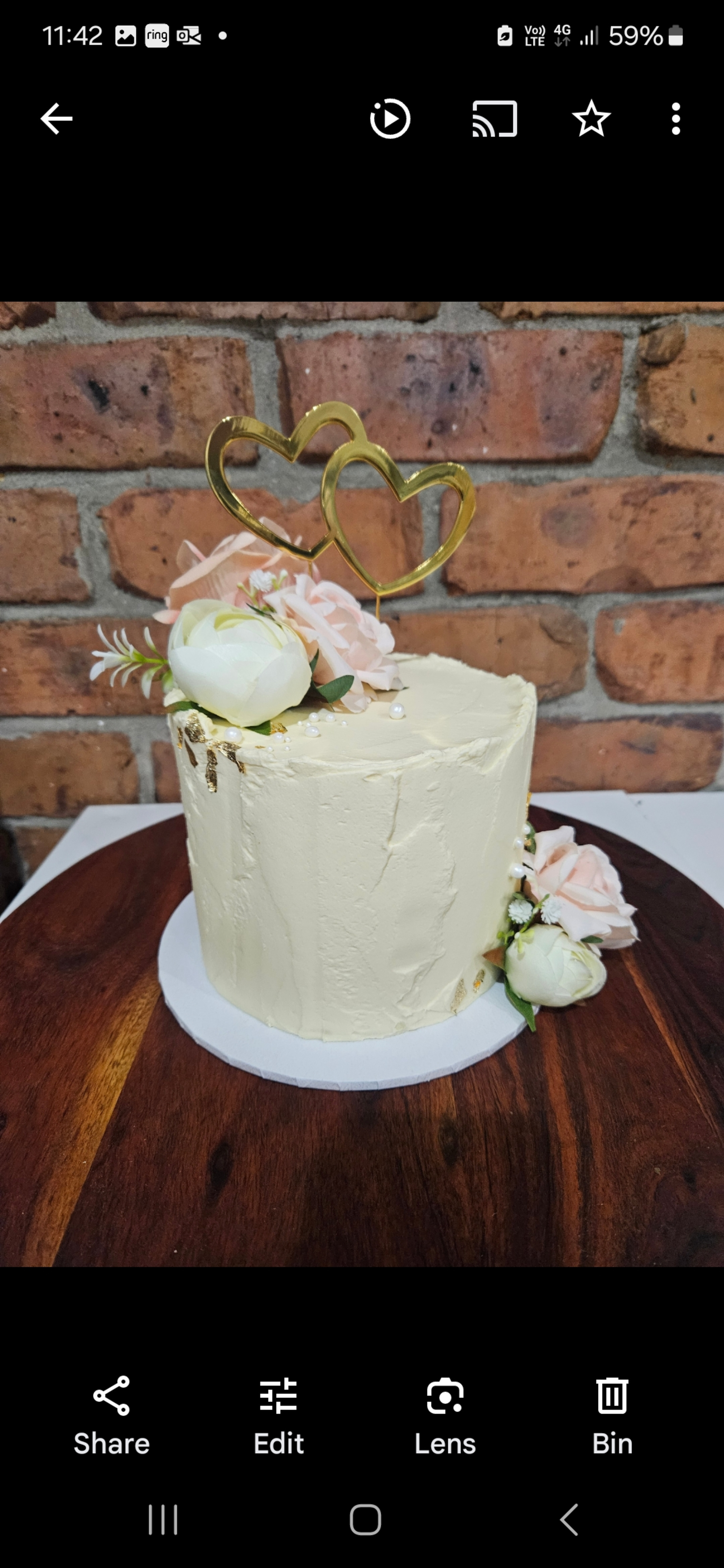 Single-tier ivory wedding cake with gold double heart topper and pastel flowers on a wooden stand.