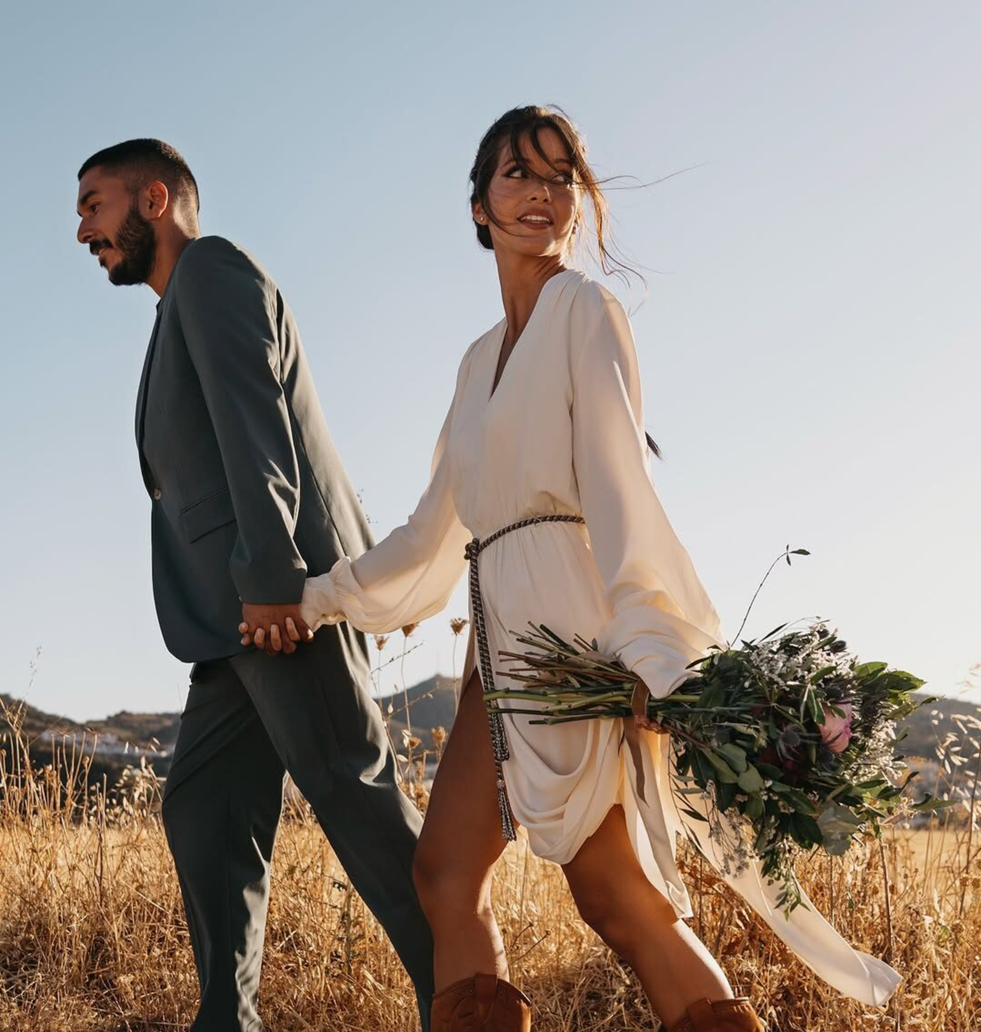 A couple walks hand in hand through a golden field at sunset, with the bride holding a lush bouquet.
