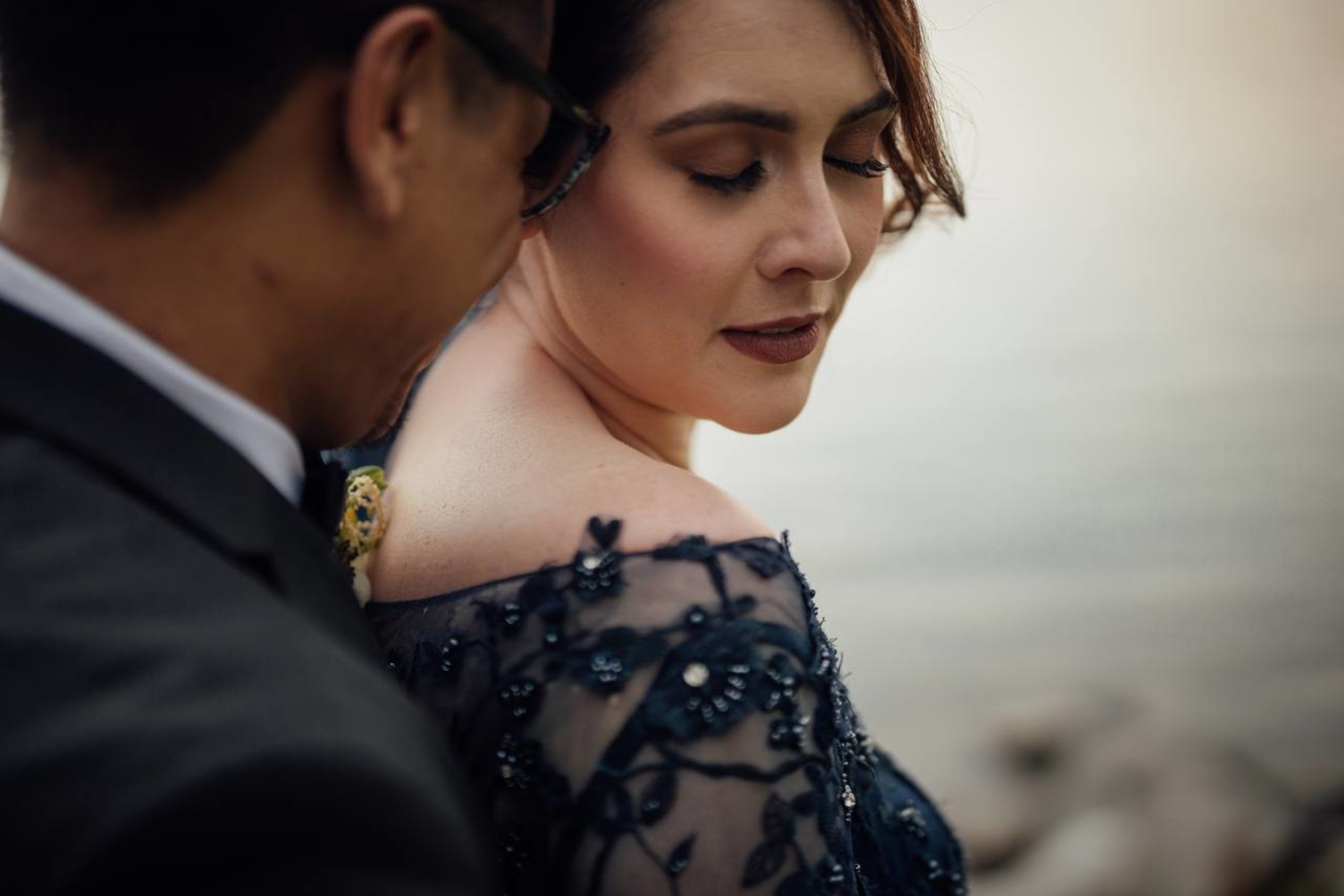 Close-up of a bride in a navy embellished dress as her partner leans in during an intimate lakeside moment.