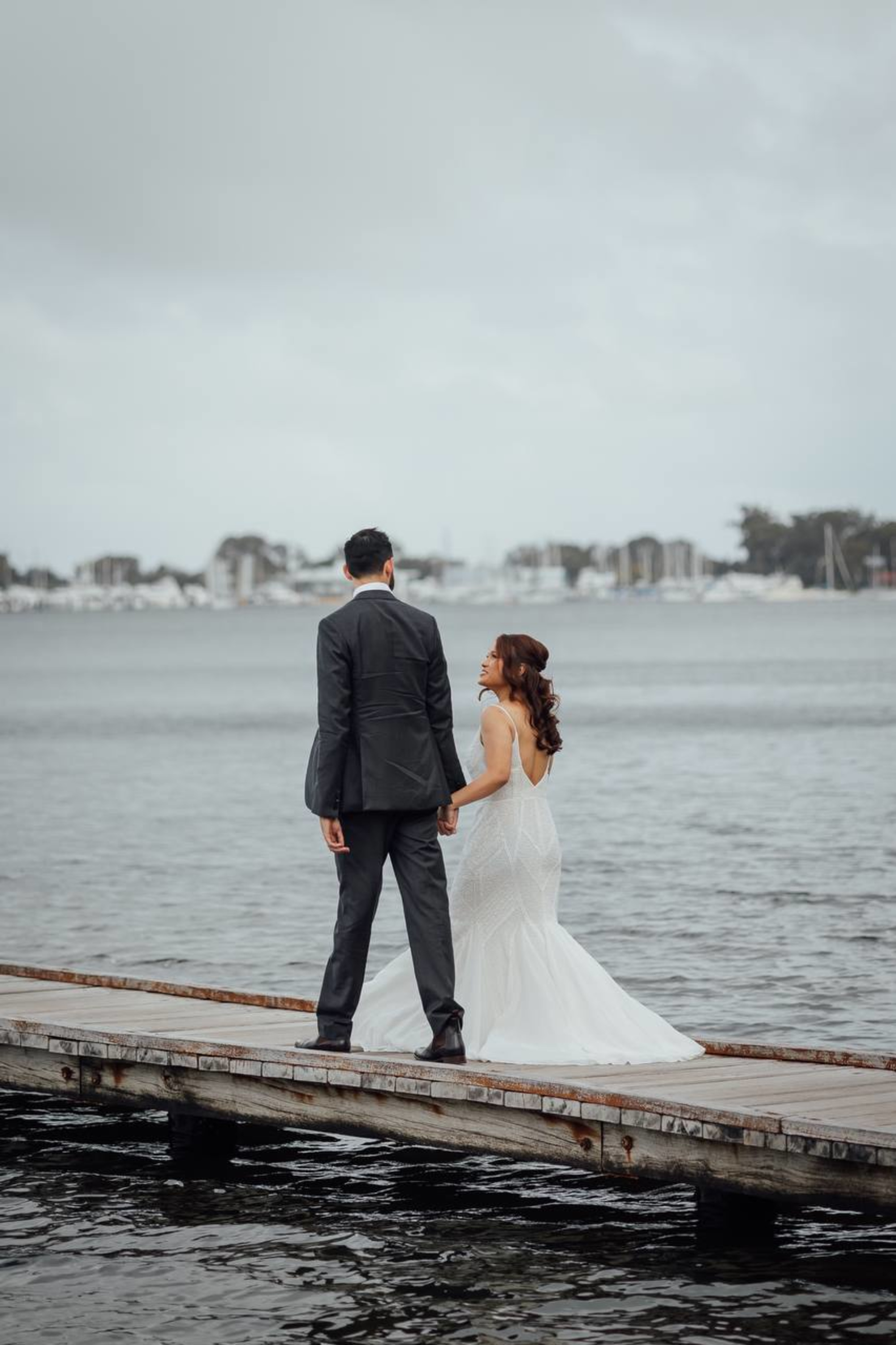 Bride and groom stand hand in hand on a dock overlooking a calm waterfront.