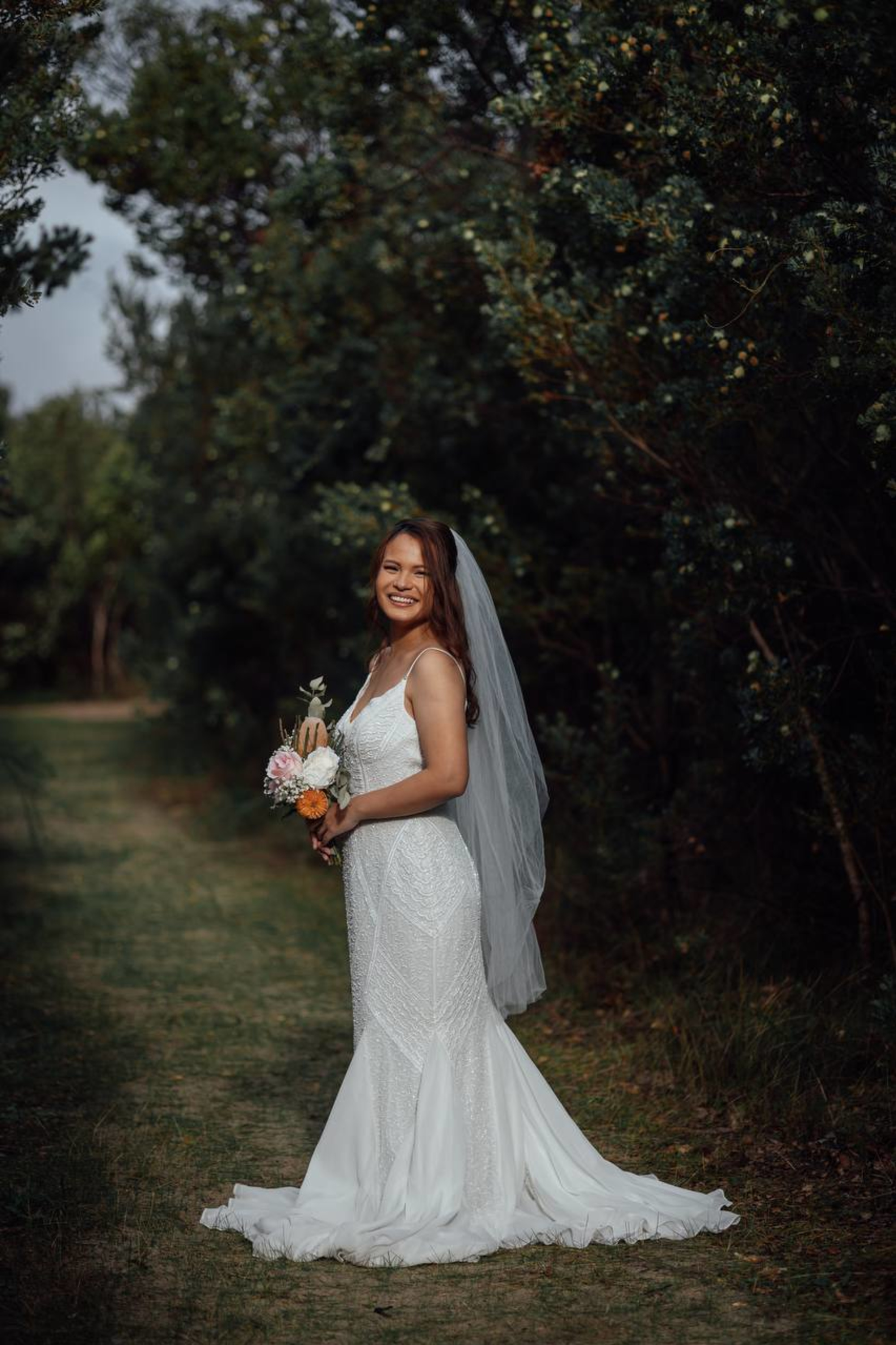 Smiling bride in a fitted white gown and veil holding a bouquet along a wooded outdoor path.