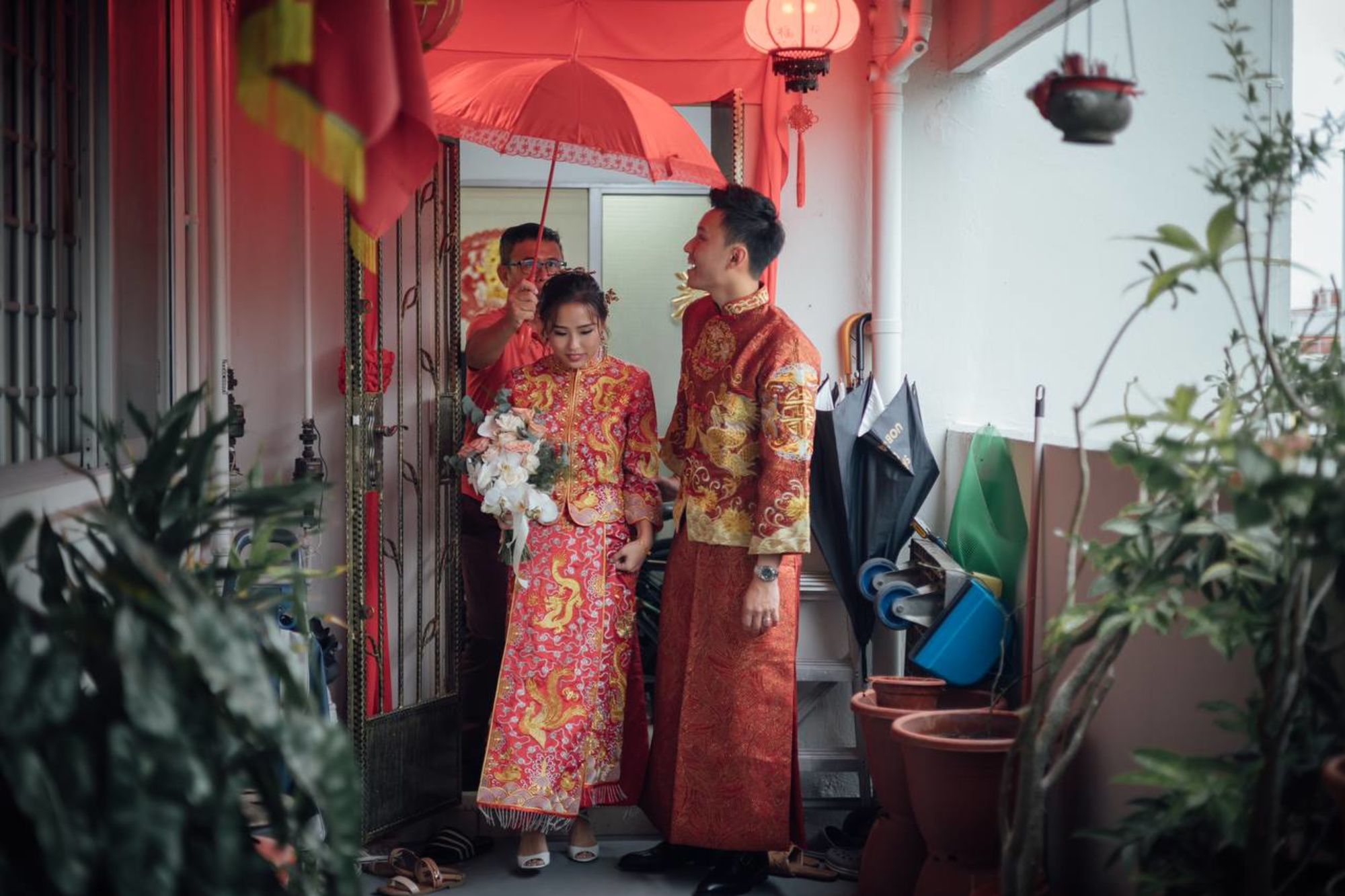 Couple in traditional Chinese wedding attire walks under a red umbrella on a balcony decorated with plants and lanterns.