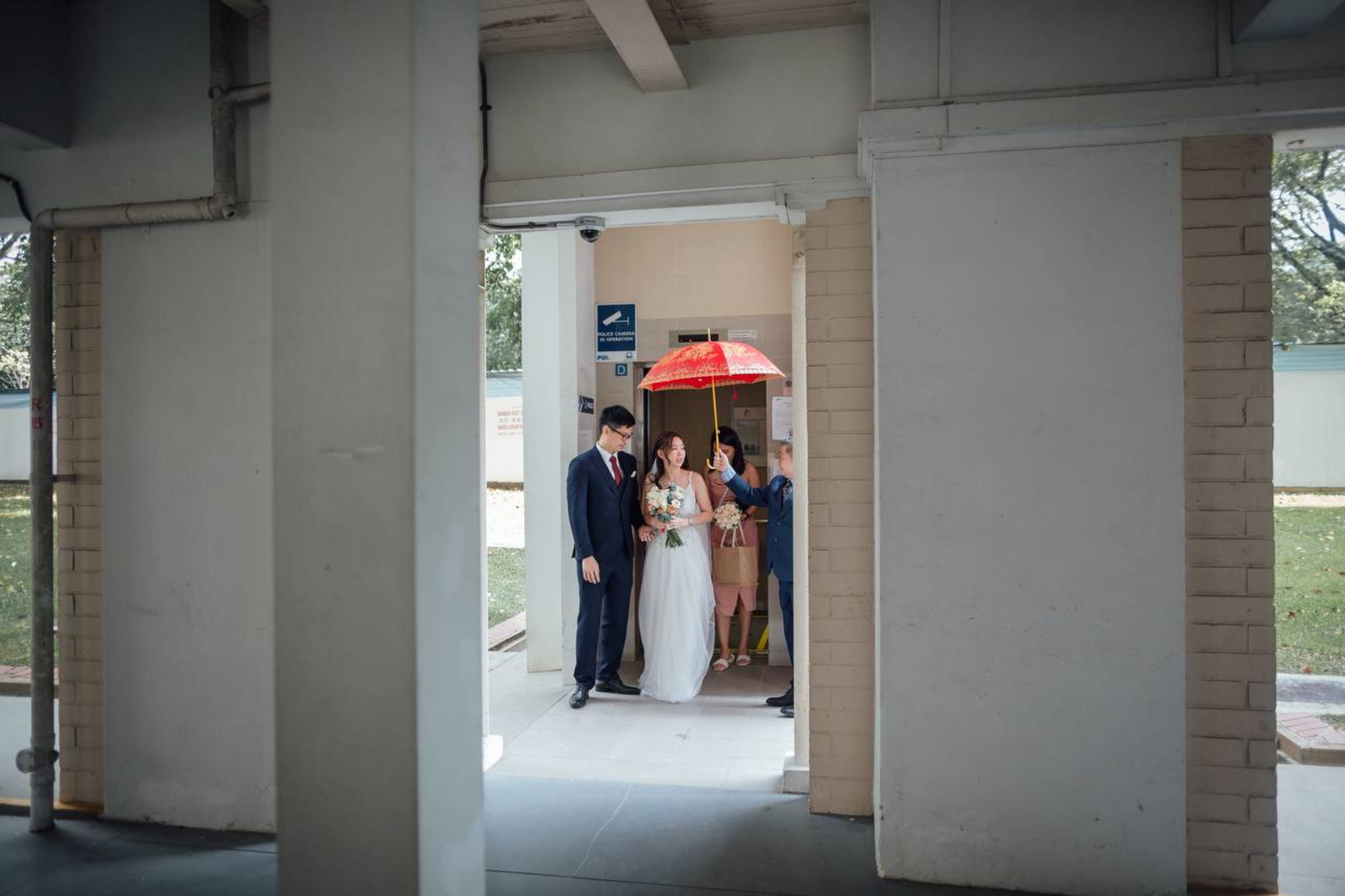 Bride and groom stand with family under a red umbrella in a corridor on their wedding day.
