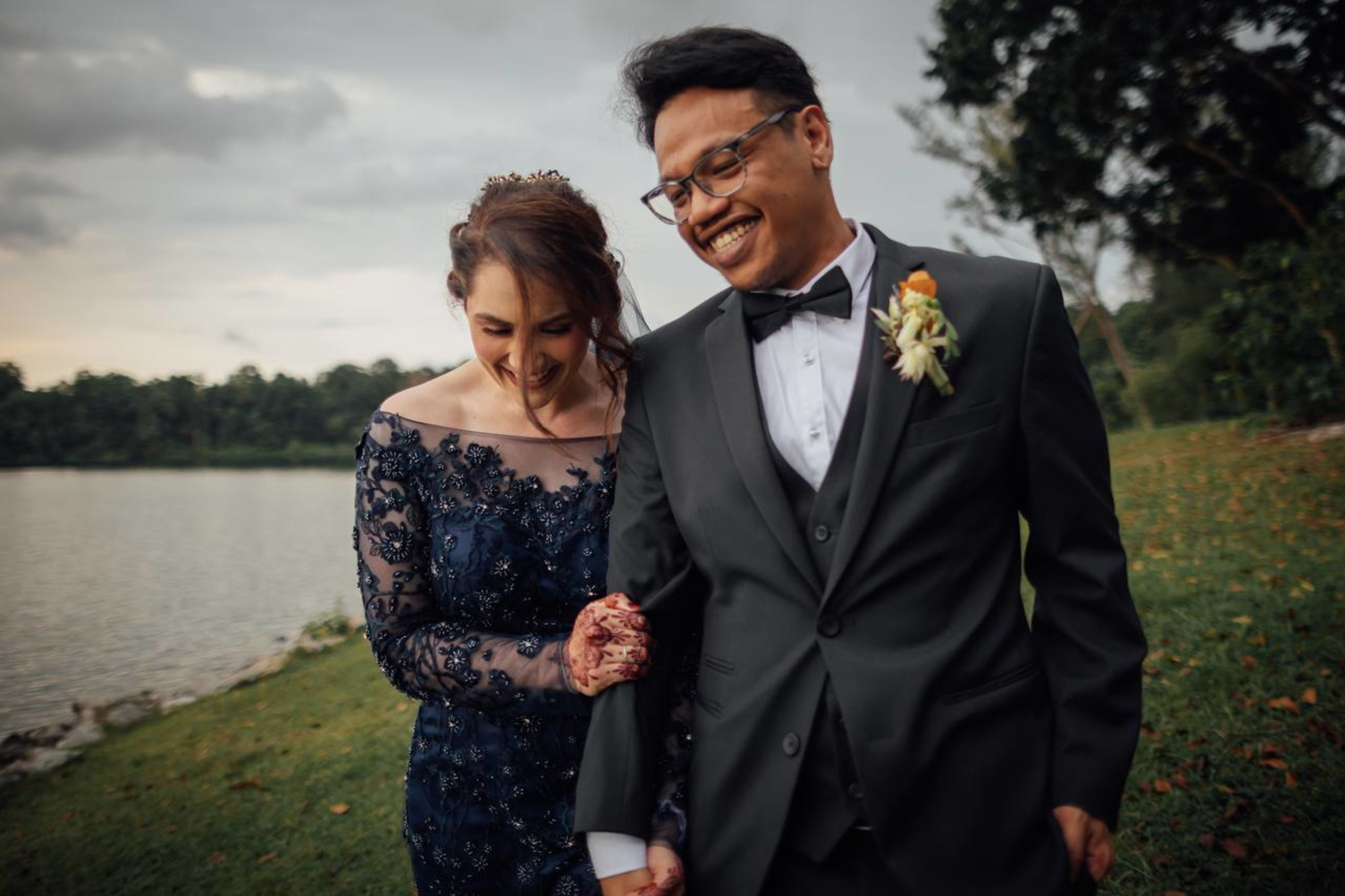 Smiling couple walks arm in arm by a lakeside lawn on their wedding day.