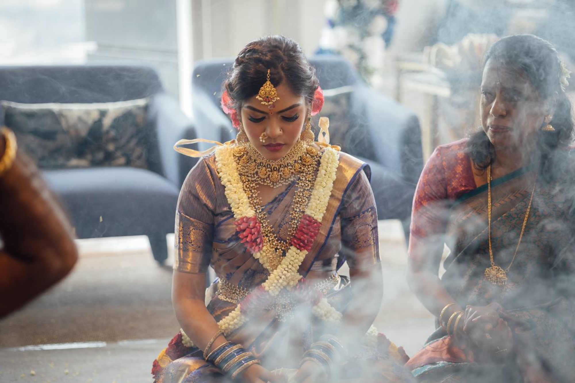 South Indian bride in traditional saree and heavy gold jewelry sits in ritual smoke during a wedding ceremony.
