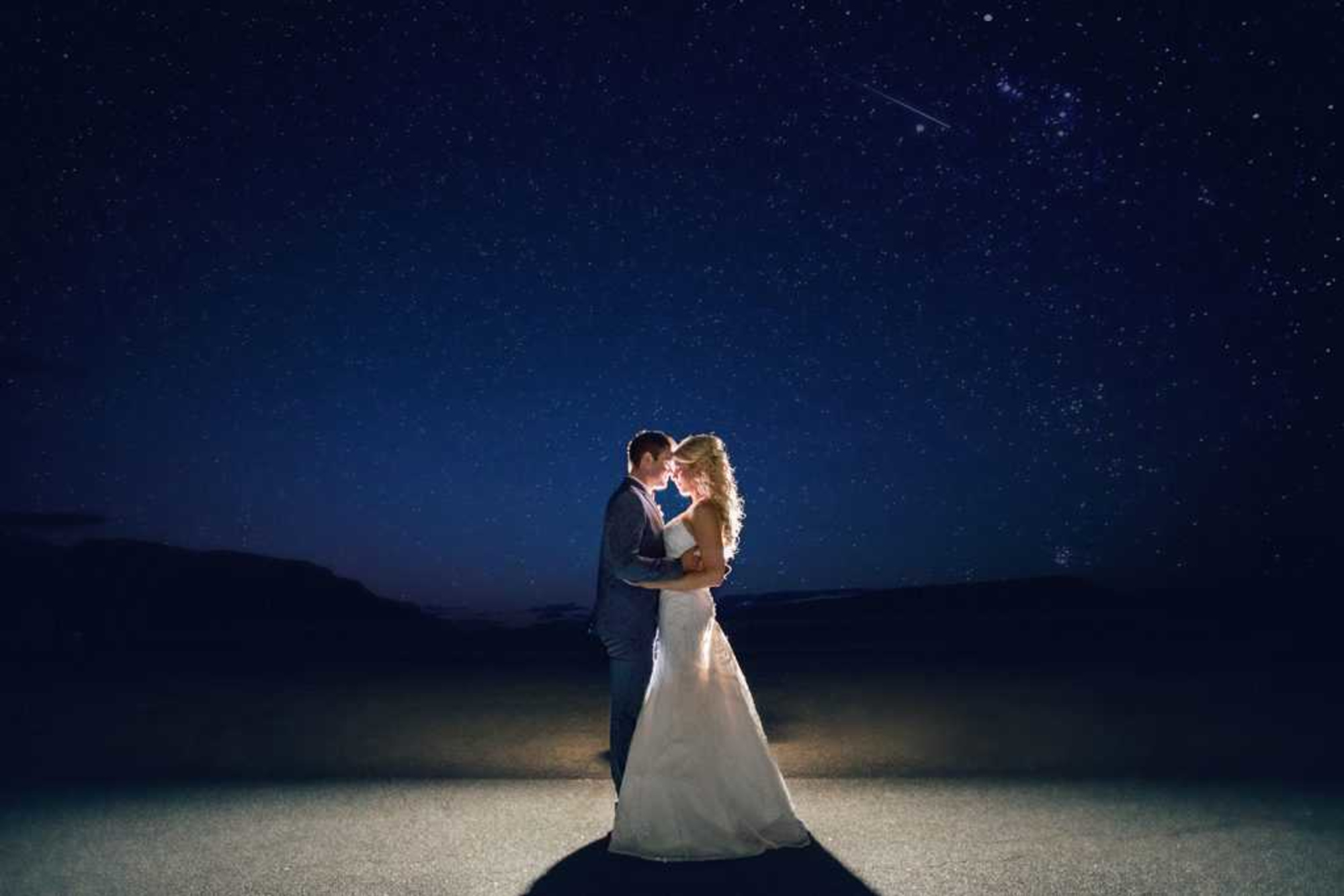 Bride and groom embrace under a starry night sky with dramatic backlighting.
